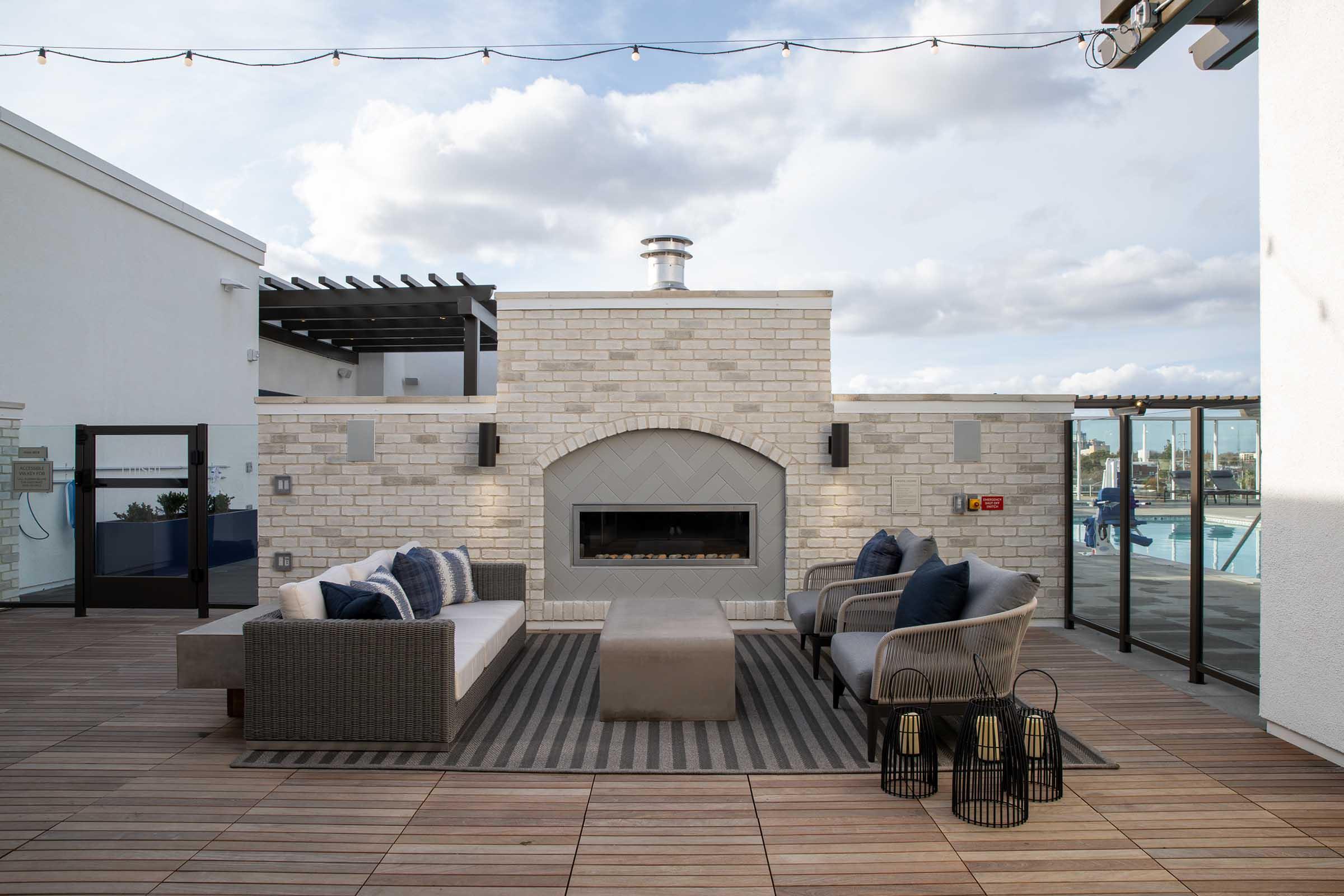 A modern outdoor lounge area featuring a stylish stone fireplace, comfortable seating with navy and white cushions, and a striped rug. Surrounding the space are glass panels and a view of a swimming pool under a partly cloudy sky, creating a relaxing atmosphere.