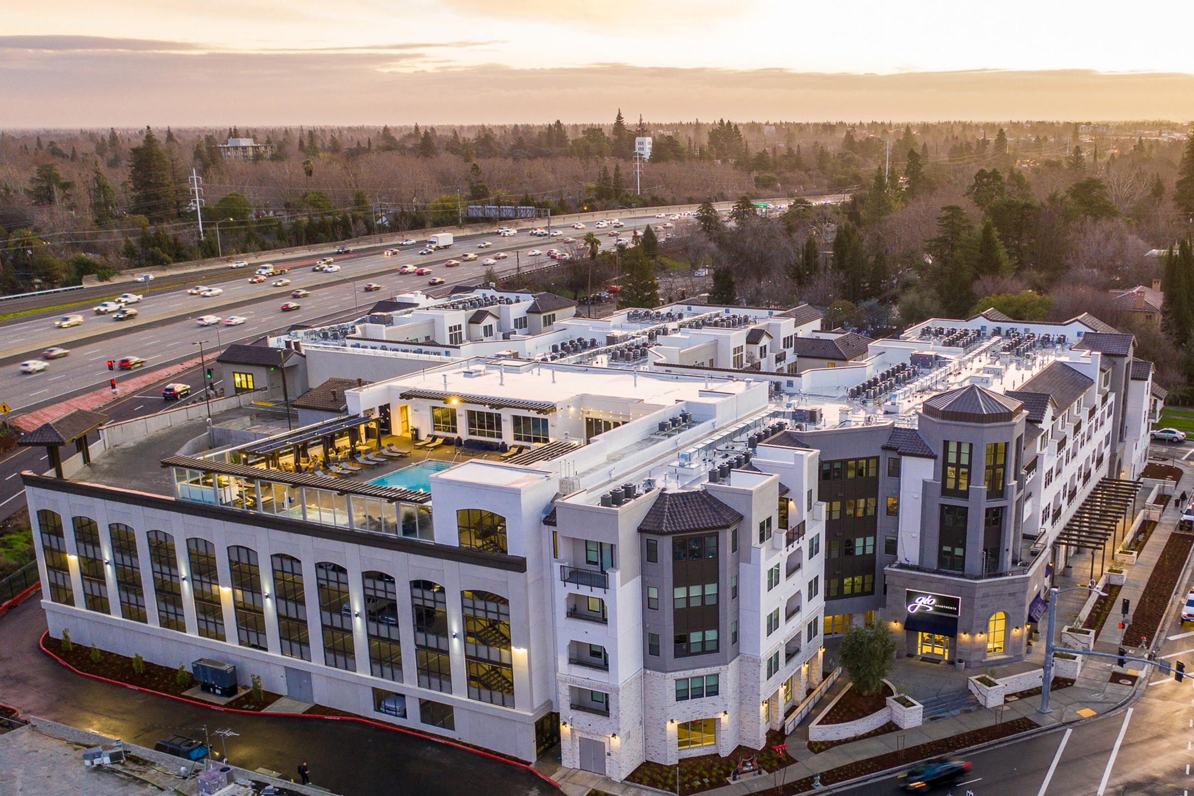 A modern multi-story hotel building with a rooftop pool and terrace, situated near a busy highway. The exterior showcases a mix of white and gray materials, with large windows and balconies. The surrounding area features trees and traffic, under a sunset sky.