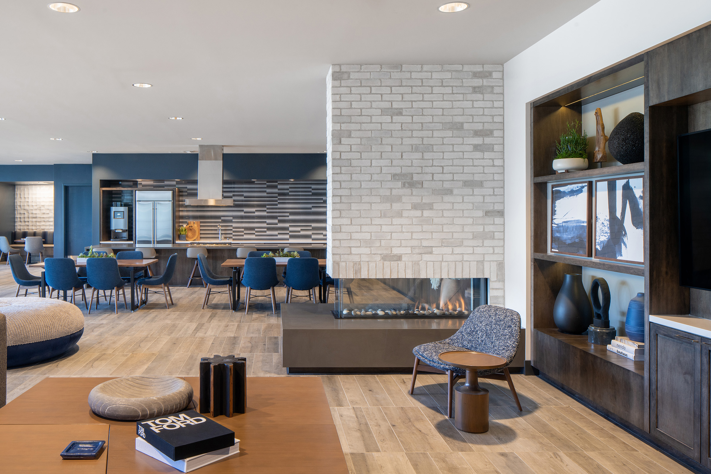 Modern interior of a stylish lounge area featuring a cozy fireplace, comfortable seating, and a mix of contemporary furniture. Large windows fill the space with natural light, while a dining area with blue chairs and a sleek kitchen is visible in the background. Decorative elements include books and art on display.