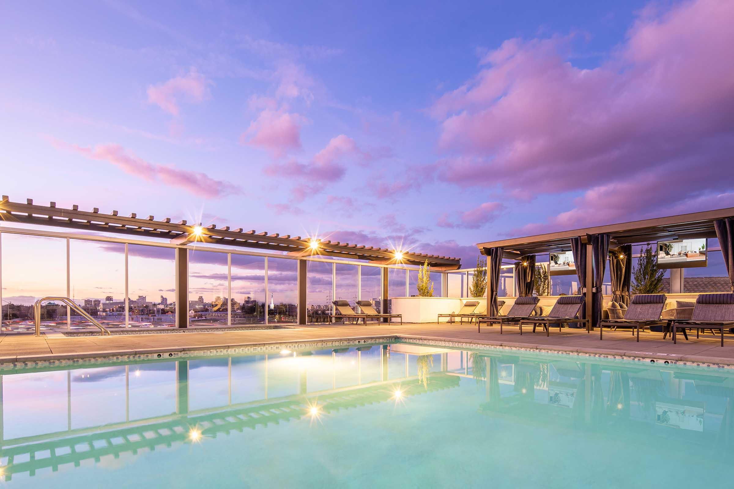 A tranquil rooftop pool at sunset, surrounded by lounge chairs and elegant cabanas. The water reflects soft pink and purple hues from the sky, with warm lights illuminating the area. Glass walls provide a panoramic view of the city skyline in the background.