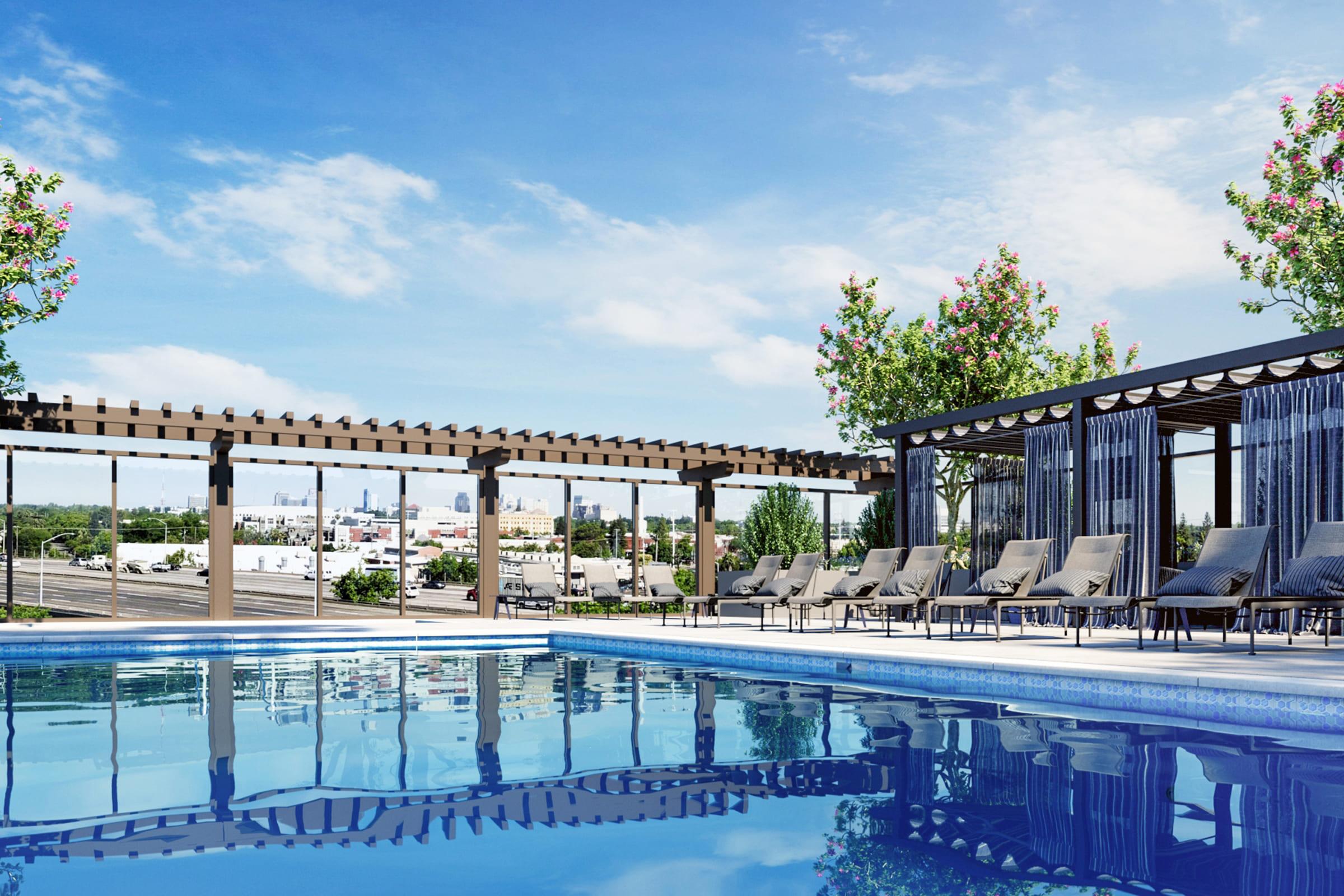 An outdoor swimming pool surrounded by lounge chairs, pergolas, and flowering trees under a blue sky. The water reflects the structures and trees, creating a serene atmosphere. In the background, buildings are visible, indicating a developed area.