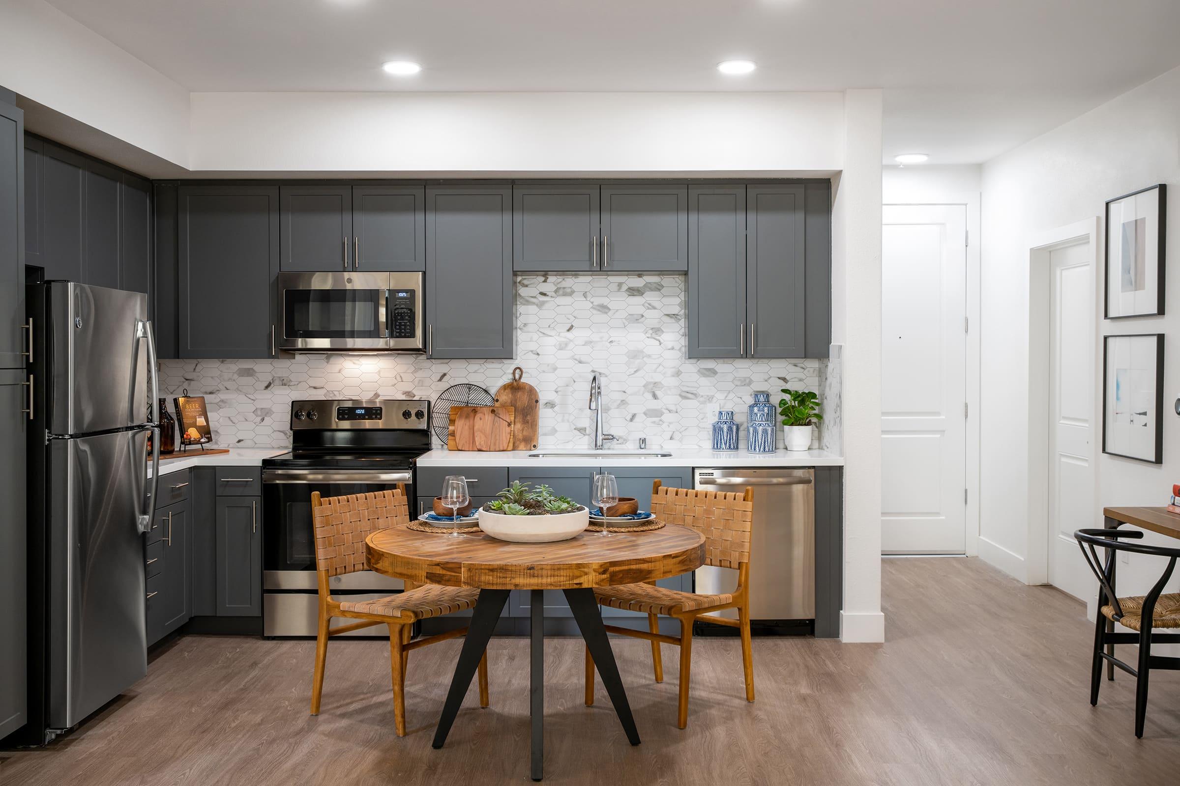 Modern kitchen featuring gray cabinets, stainless steel appliances, and a stylish marble backsplash. A round wooden dining table with two chairs is set nearby, complemented by decorative items on the countertop. Well-lit space with simple artwork and minimalistic decor gives a welcoming atmosphere.