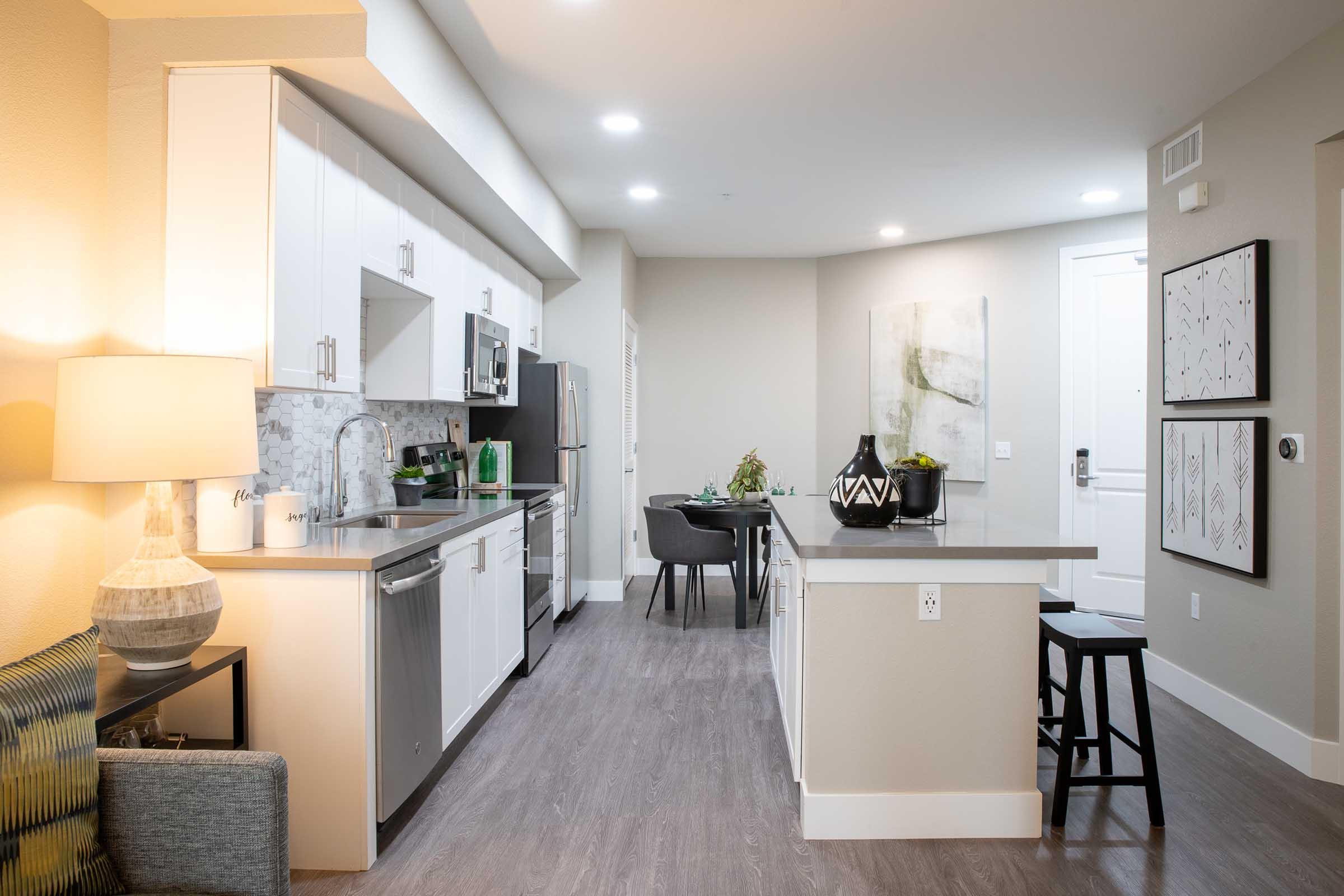 Modern kitchen and dining area featuring sleek white cabinetry, stainless steel appliances, and a gray countertop. The space is well-lit with overhead lighting and has contemporary decor, including a table with chairs and abstract wall art. A cozy seating area is visible in the foreground.
