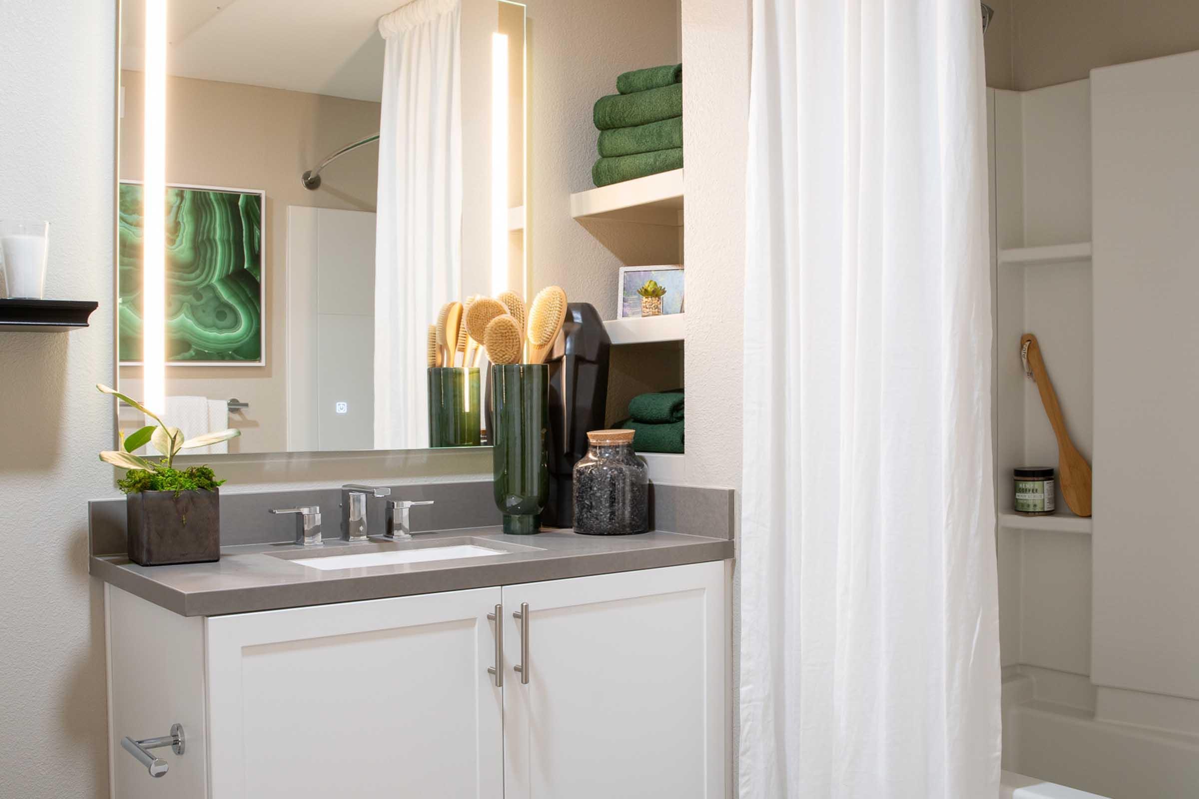 A modern bathroom featuring a sleek gray countertop with dual faucets, a large mirror, and a decorative plant. Closed white curtains separate the shower area, while green towels are neatly stacked on a shelf. Artwork with green tones hangs on the wall, adding a touch of elegance to the space.