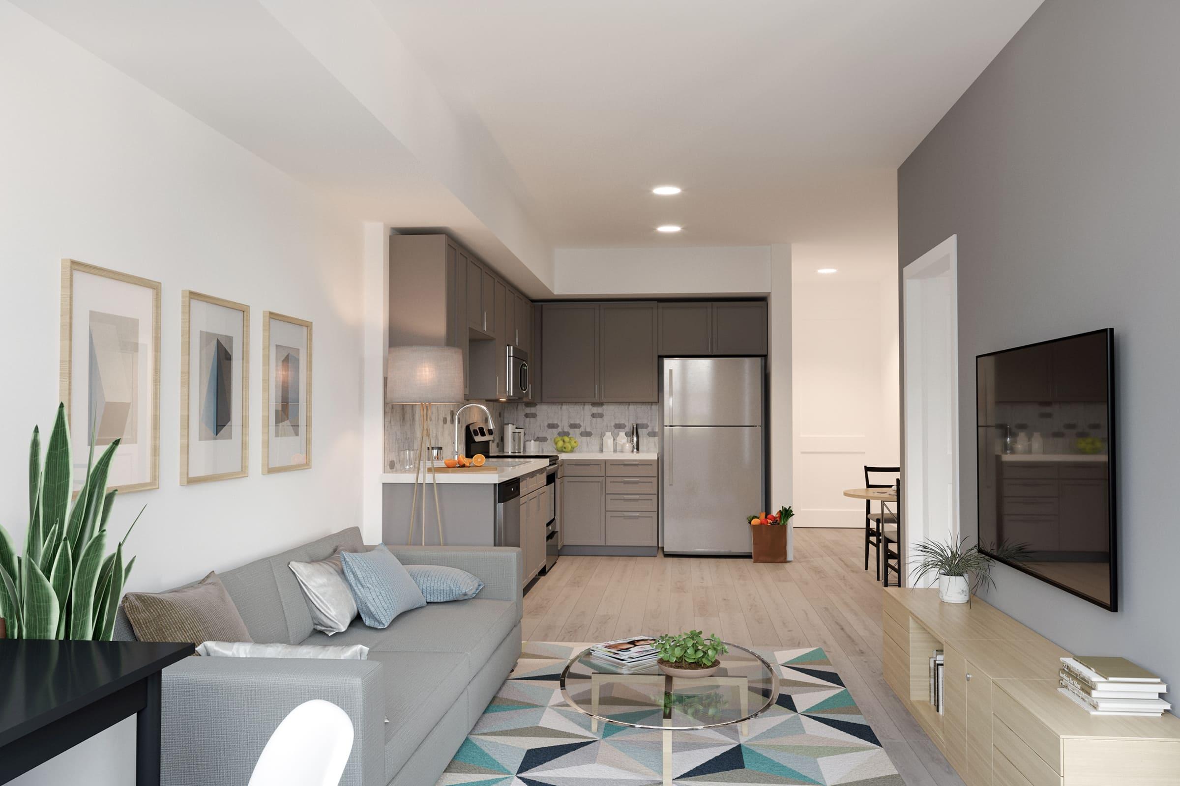 A modern living room and kitchen area featuring a gray sofa with decorative pillows, a round coffee table with plants, and framed artworks on the wall. The kitchen includes gray cabinets, stainless steel appliances, and a dining table in the background, with natural light streaming in through the windows.