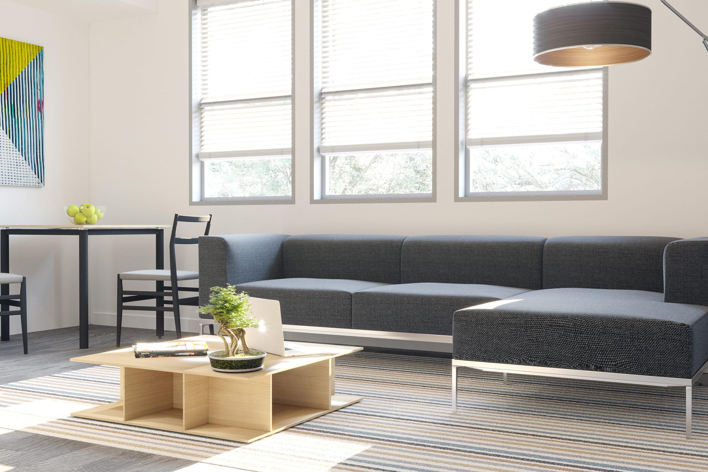 A bright and modern living room featuring a gray sectional sofa, a minimalist wooden coffee table with a plant, and a laptop on it. Light streams in through large windows covered with blinds. A small dining table with black chairs is visible in the background, and a bowl of green apples sits on the table.