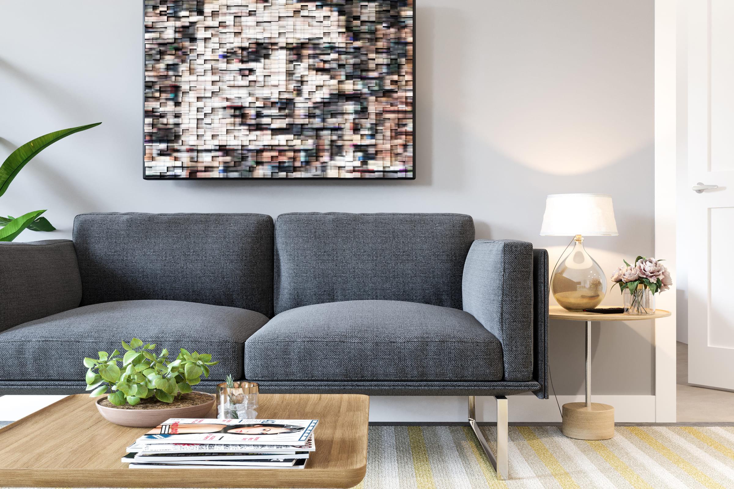 A modern living room featuring a gray couch with a textured surface, a small side table with a lamp and flowers, and a wooden coffee table with magazines. A colorful abstract artwork hangs on the wall above the couch, and there's a green plant near the couch, complementing the cozy atmosphere.