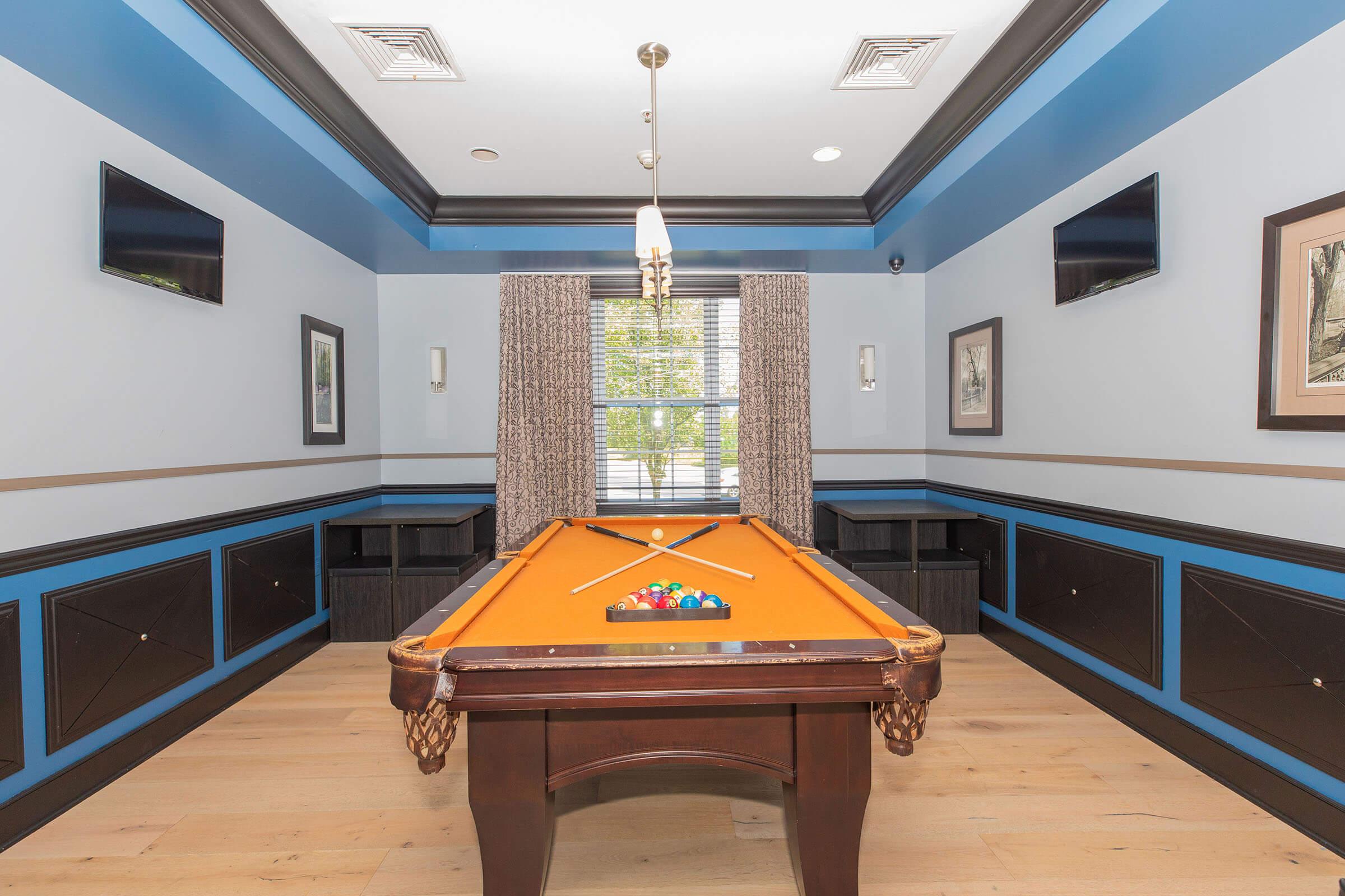 View of a game room featuring a billiards table with colorful balls arranged on it. The room has light-colored wooden flooring, blue and gray walls with decorative molding, and two wall-mounted TVs. A large window with patterned curtains allows natural light to enter, enhancing the inviting atmosphere.