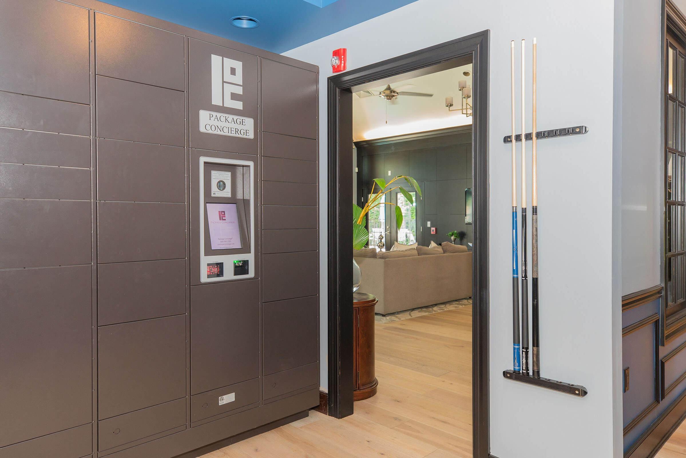 A modern package concierge locker situated in a hallway, with a doorway leading to a living area featuring a couch and plants. A pool table with cues is visible on the right side, and the decor has a contemporary style with light-colored flooring and blue ceiling accents.