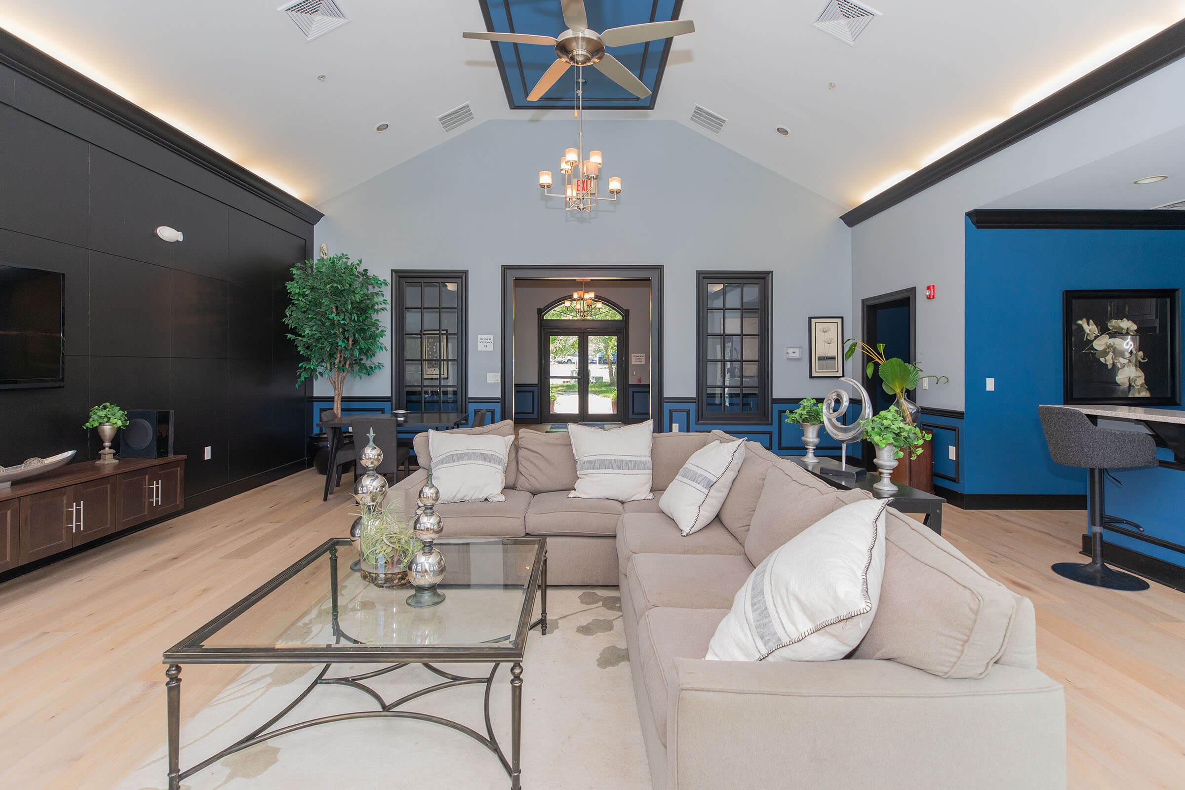 A modern lounge area featuring a large beige sectional sofa around a glass coffee table. There are decorative vases on the table and lush green plants in the corners. The walls are painted in shades of blue and black, with a chandelier hanging from the ceiling, creating an inviting atmosphere.