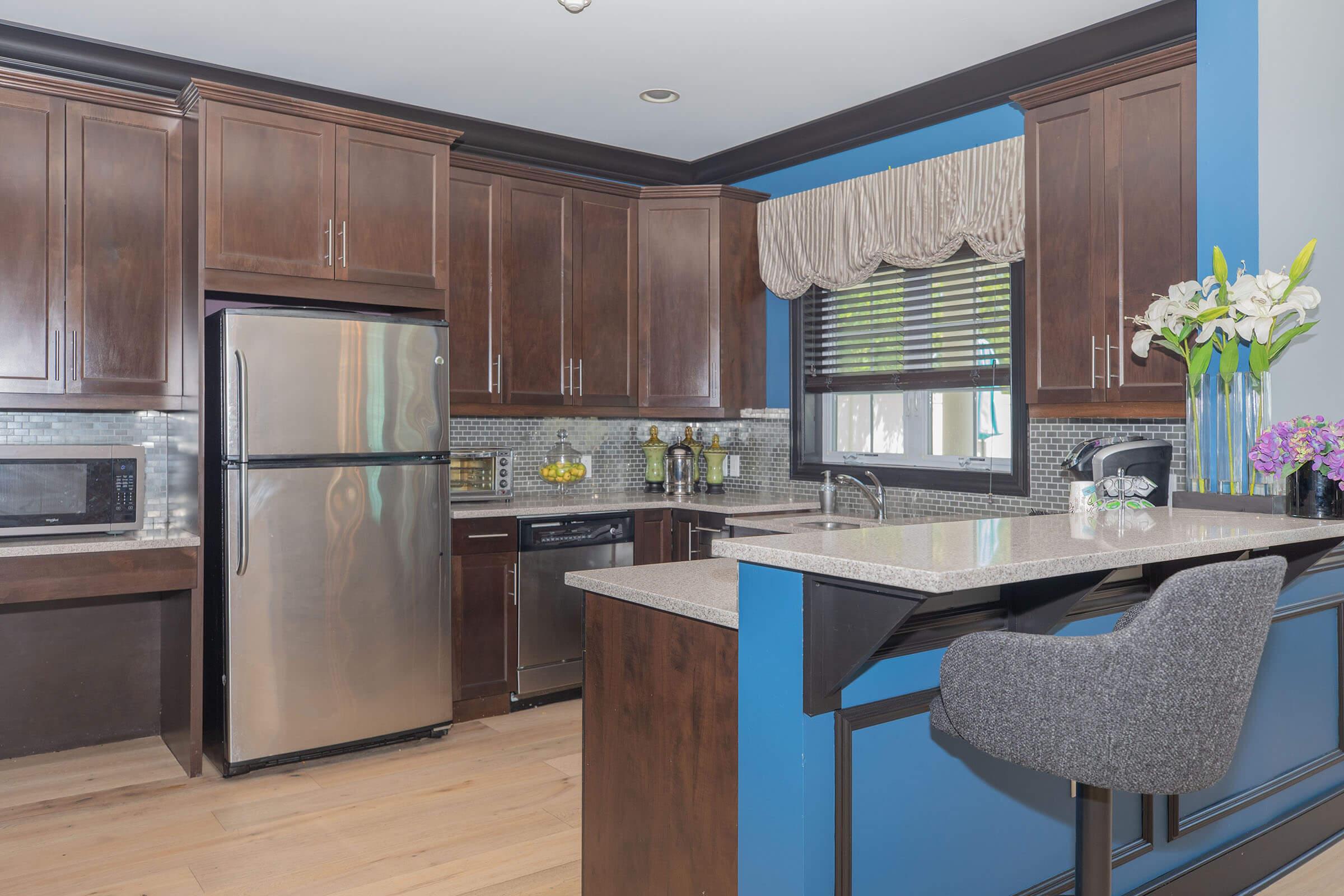 Modern kitchen featuring dark wood cabinets, stainless steel appliances including a refrigerator and dishwasher, a granite countertop with a bar area, and a window with blinds. Decorative elements include a vase of flowers and kitchen utensils, with a light blue accent wall contrasting with the dark cabinetry.