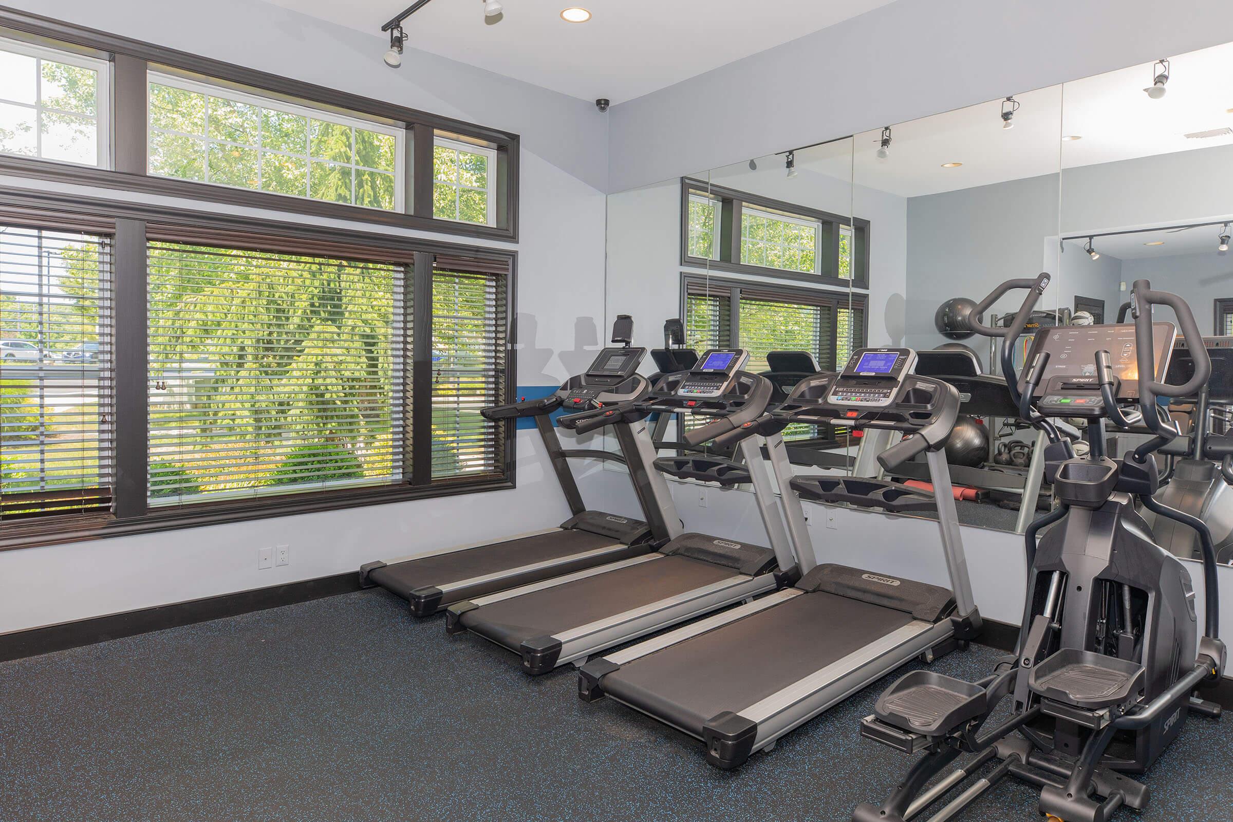 A well-lit gym room featuring three treadmills lined up against a window with views of greenery. Mirrors on the walls enhance the space, and additional gym equipment appears in the background, creating an inviting atmosphere for workouts.