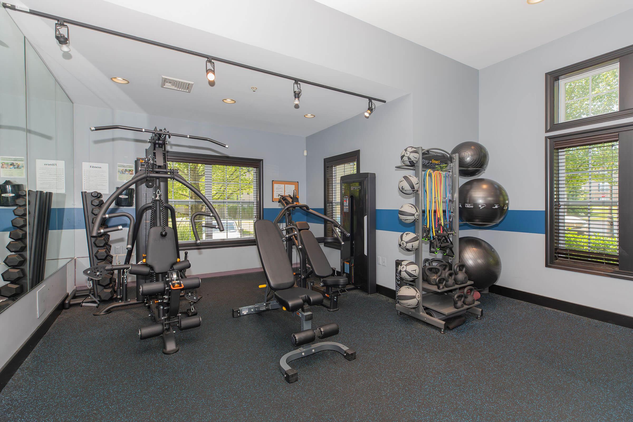 A modern gym interior featuring a multi-station exercise machine, a bench, and a selection of weight plates. The walls are painted in light colors with blue accents, and large windows allow natural light to enter. Additionally, there are exercise balls and resistance bands organized on a rack.