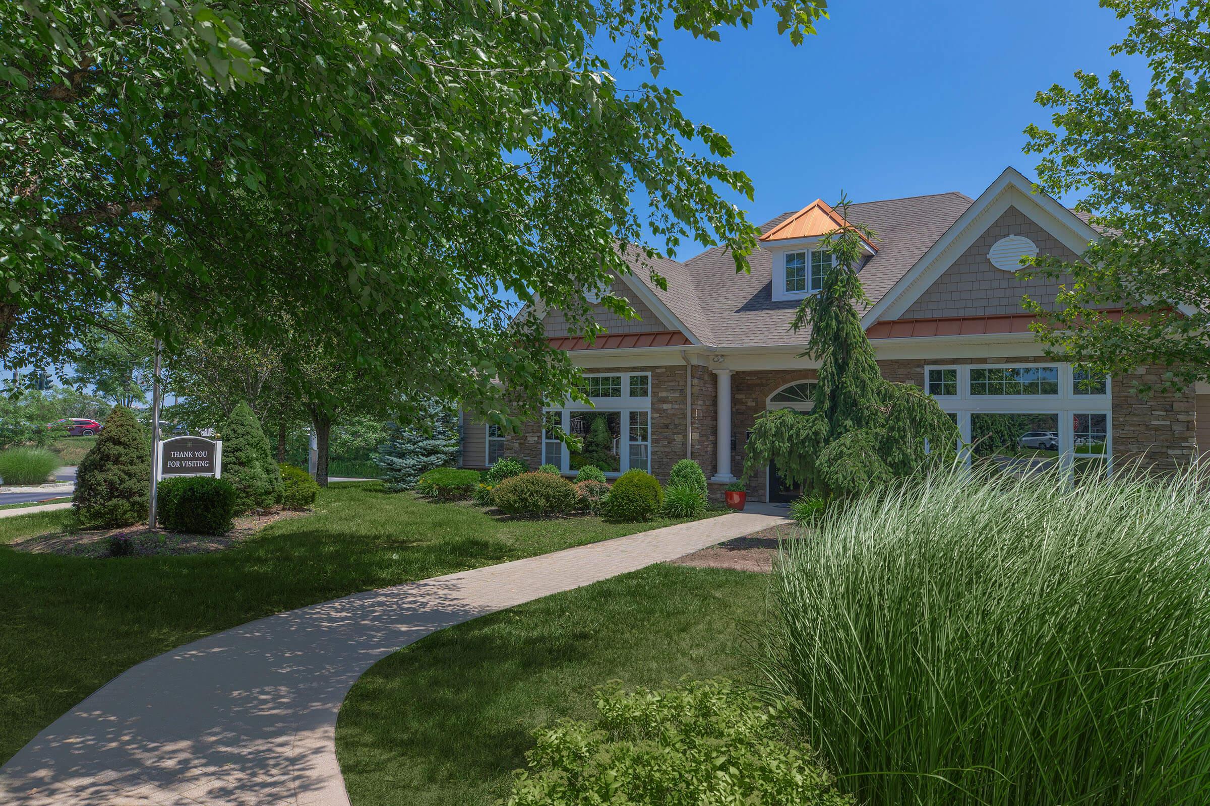 A picturesque suburban home surrounded by lush greenery and well-maintained landscaping. The house features a stone exterior, a welcoming front walkway, and large windows, with a sign at the entrance and tall grass swaying gently in the breeze under a clear blue sky.