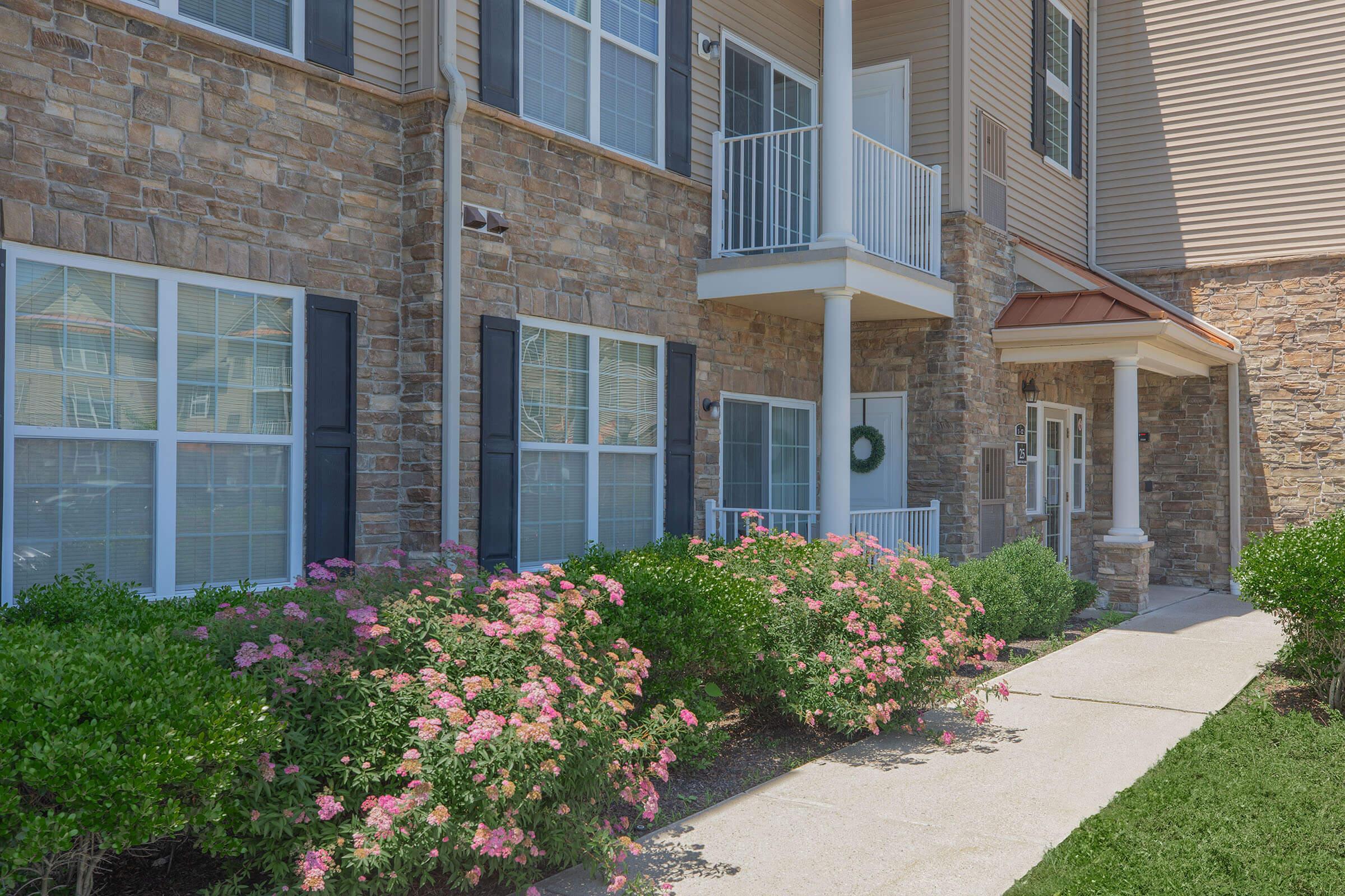 A residential building with a stone and siding exterior. The entrance features a porch with white railings and a roof overhang. Flowering shrubs with pink blossoms frame a concrete walkway leading to the front door, which is adorned with a wreath. Sunlight brightens the scene.