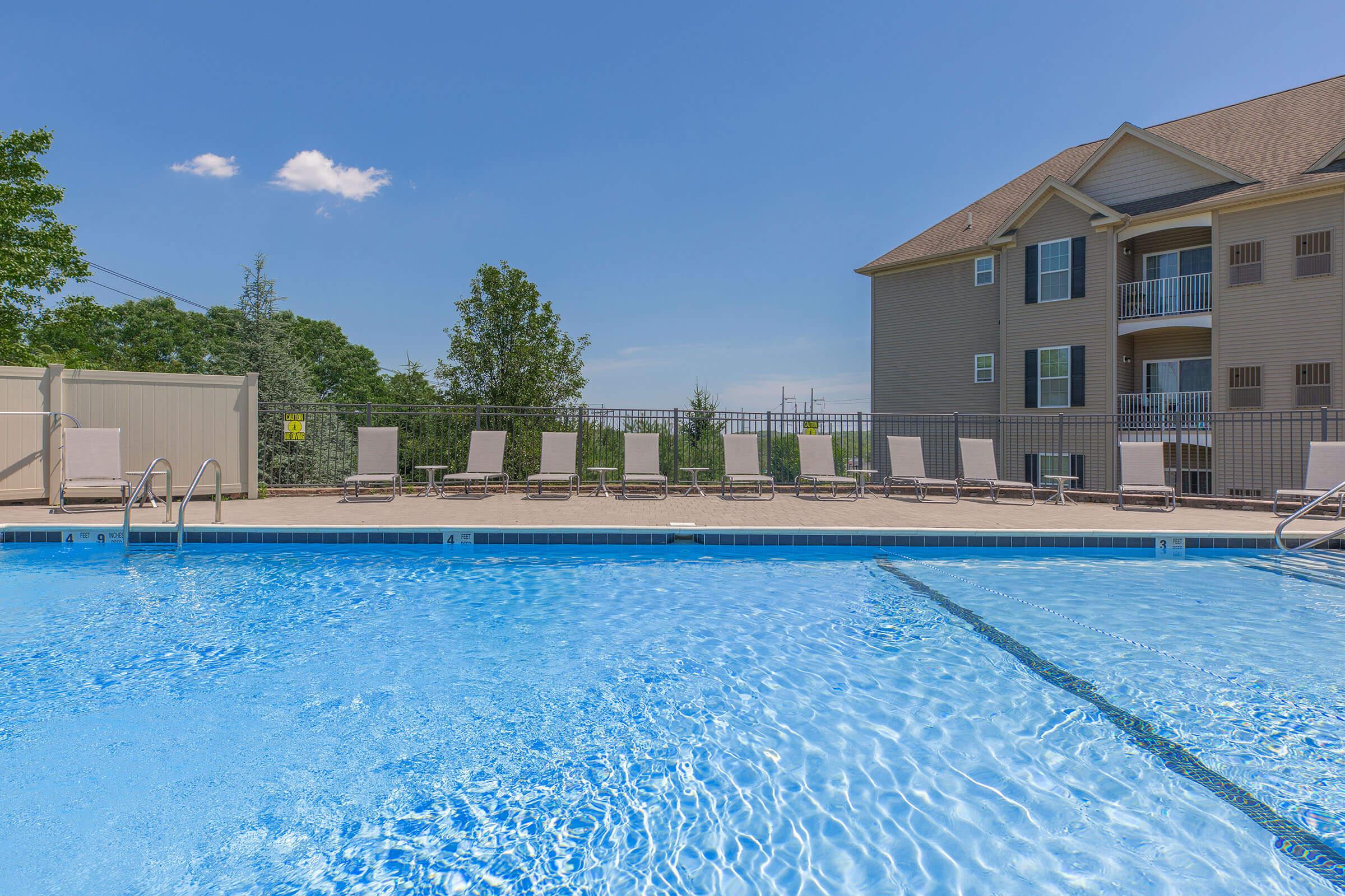 A clear swimming pool surrounded by lounge chairs under a sunny sky, with a residential building in the background. The pool features a smooth surface reflecting the blue sky, and the area is enclosed with a fence, providing a calm and inviting atmosphere for relaxation.