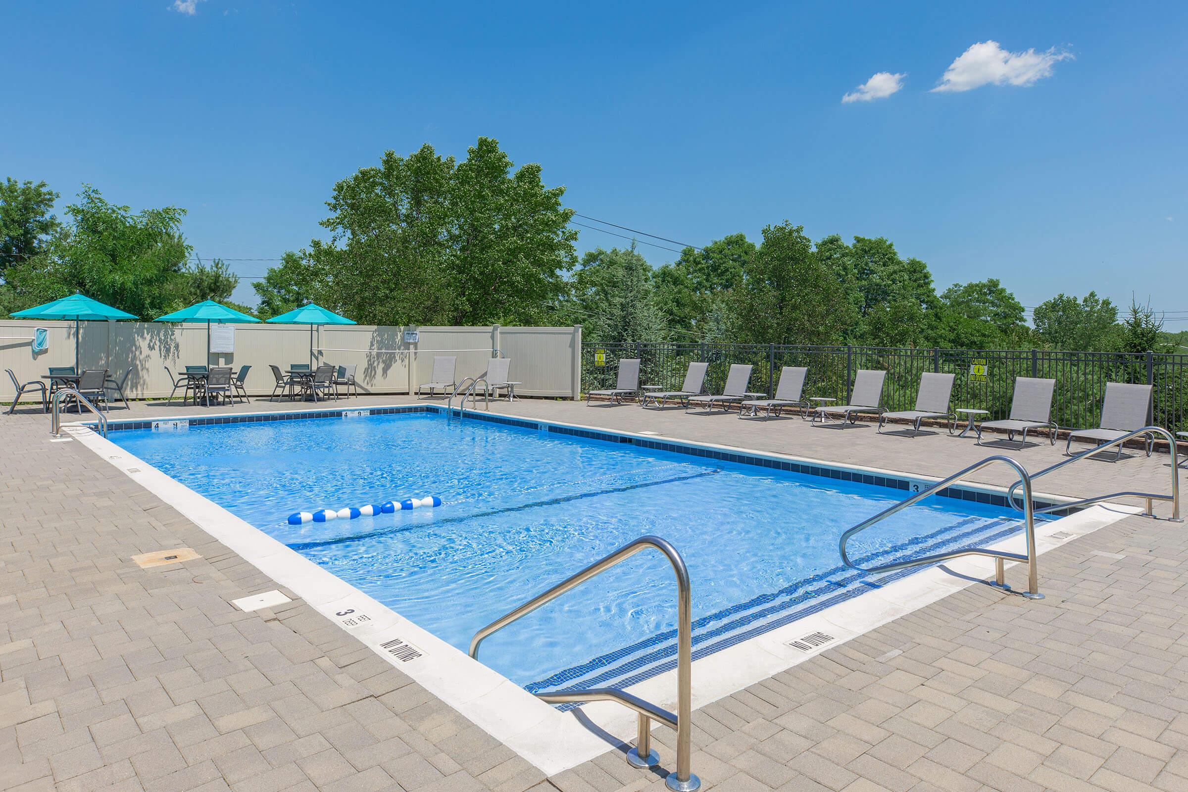 A clear blue swimming pool surrounded by lounge chairs and green trees under a bright blue sky with a few fluffy clouds. Sunshades are set up near the poolside, creating a relaxing outdoor space.