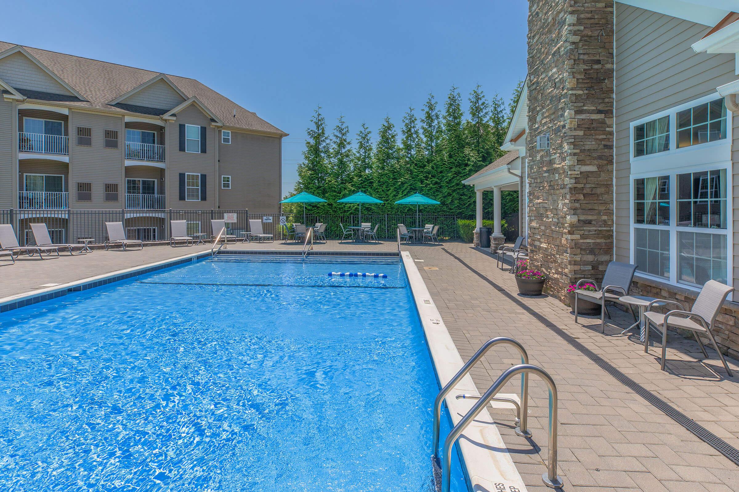 A clear blue swimming pool surrounded by a sunlit patio. Lounge chairs line the poolside, and green umbrellas provide shade. In the background, there are modern apartment buildings with balconies. Lush greenery adds to the relaxing atmosphere. Perfect for leisure and relaxation on a sunny day.
