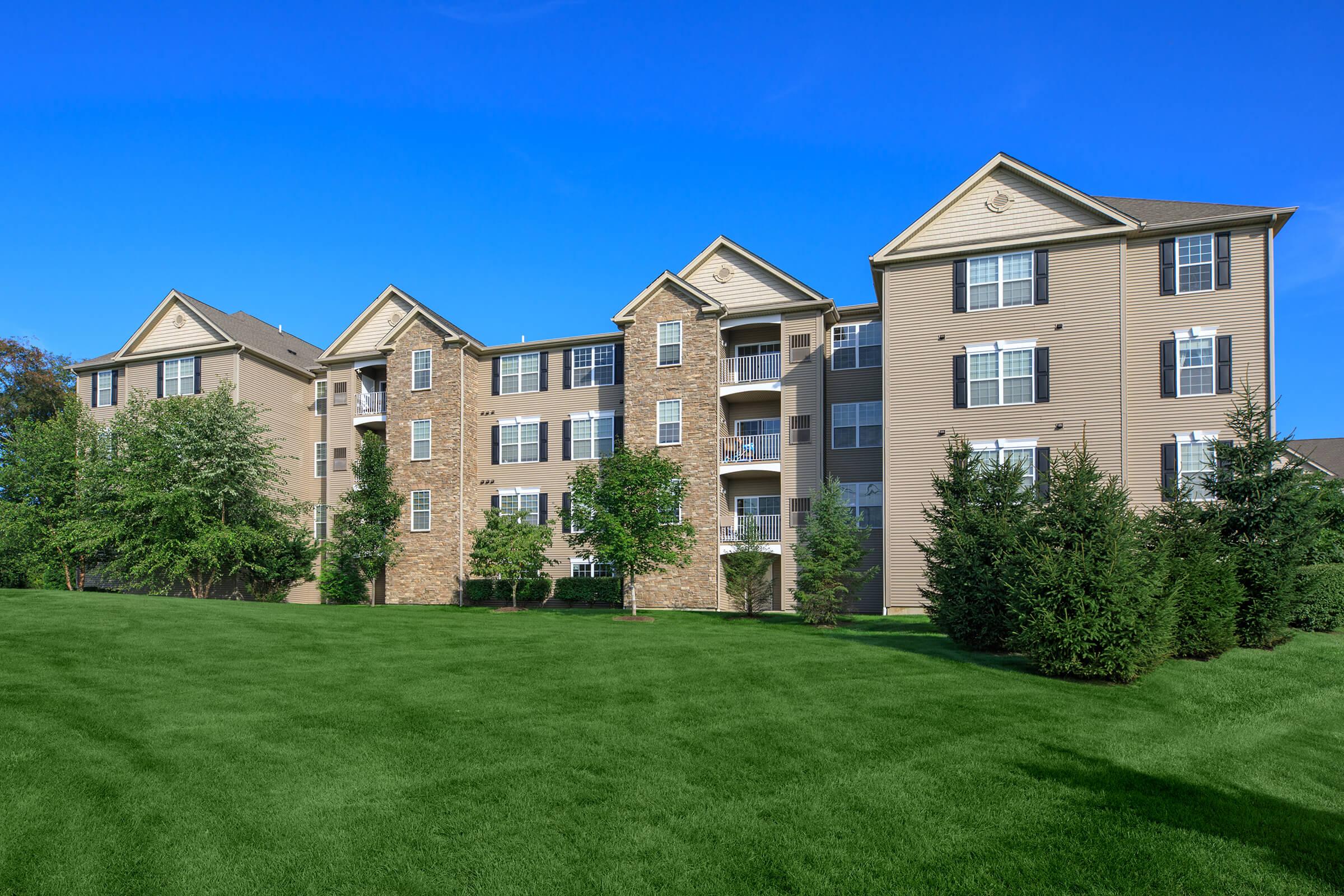 A modern multi-story apartment building featuring a mix of brick and siding exteriors, surrounded by well-maintained lawns and shrubbery under a clear blue sky.