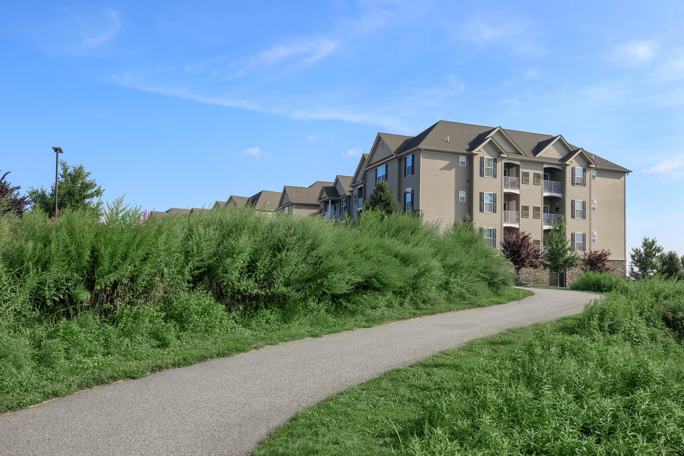 A paved pathway leading through tall green grass towards a row of modern apartment buildings under a clear blue sky. The buildings feature multiple balconies and are surrounded by landscaped greenery.