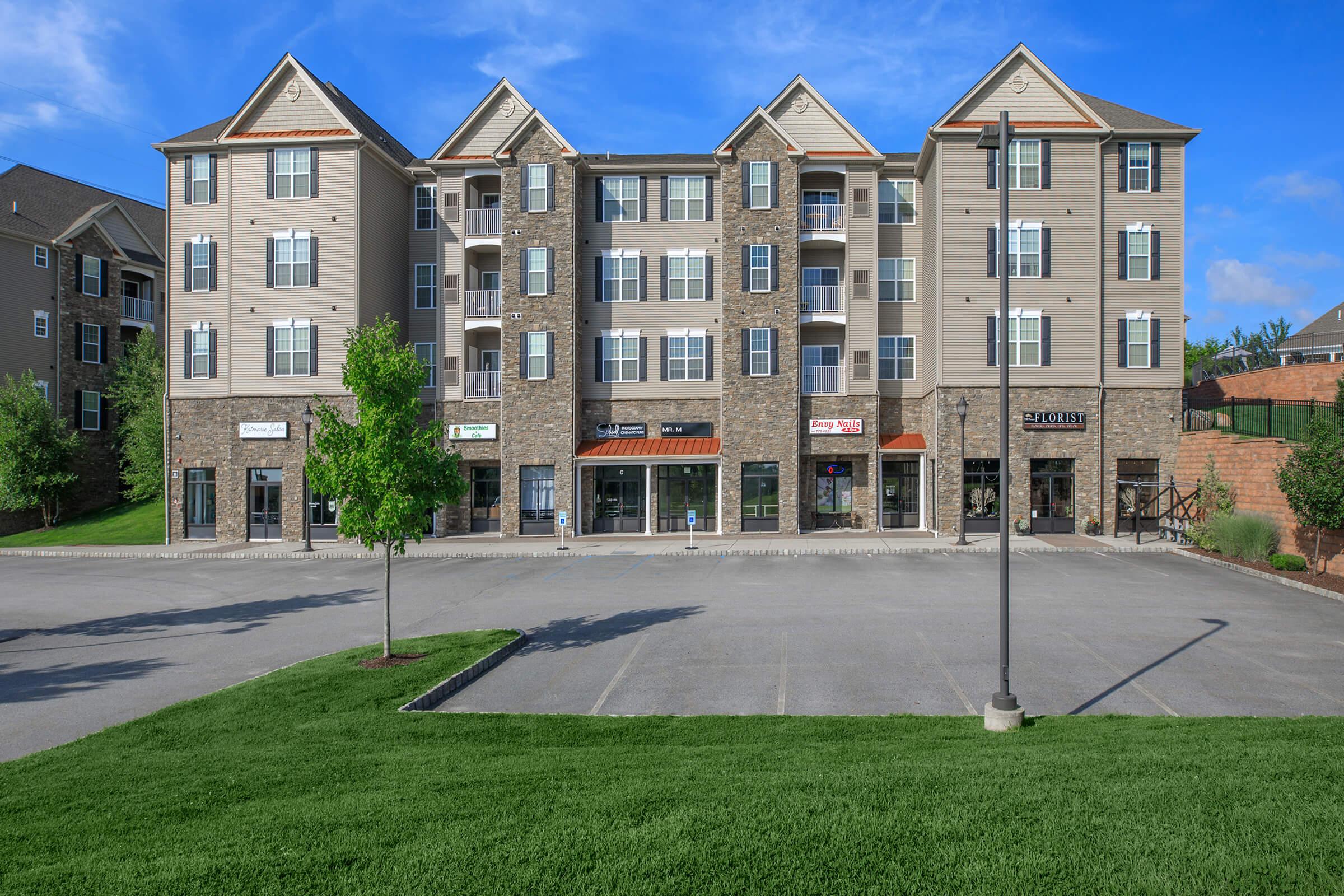 A modern three-story commercial building featuring a combination of stone and siding exteriors, large windows, and several storefronts on the ground level. The surrounding area includes a well-maintained green lawn and a clear blue sky, indicating a pleasant day.