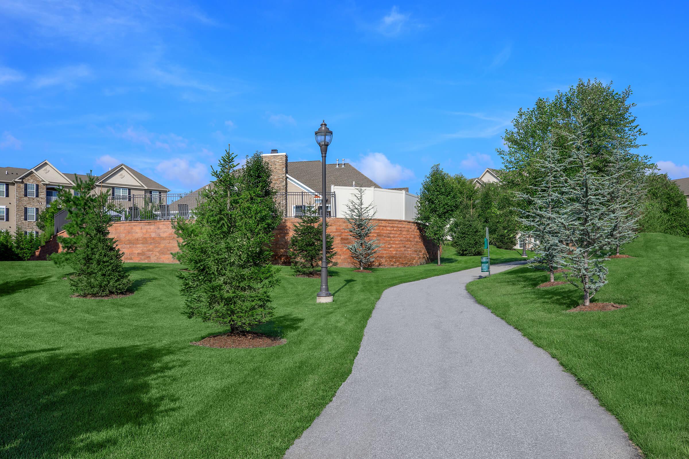 A peaceful park scene featuring a winding pathway surrounded by neatly maintained green grass and young trees. In the background, a residential area with modern houses is visible, and a lamp post stands alongside the path under a clear blue sky.