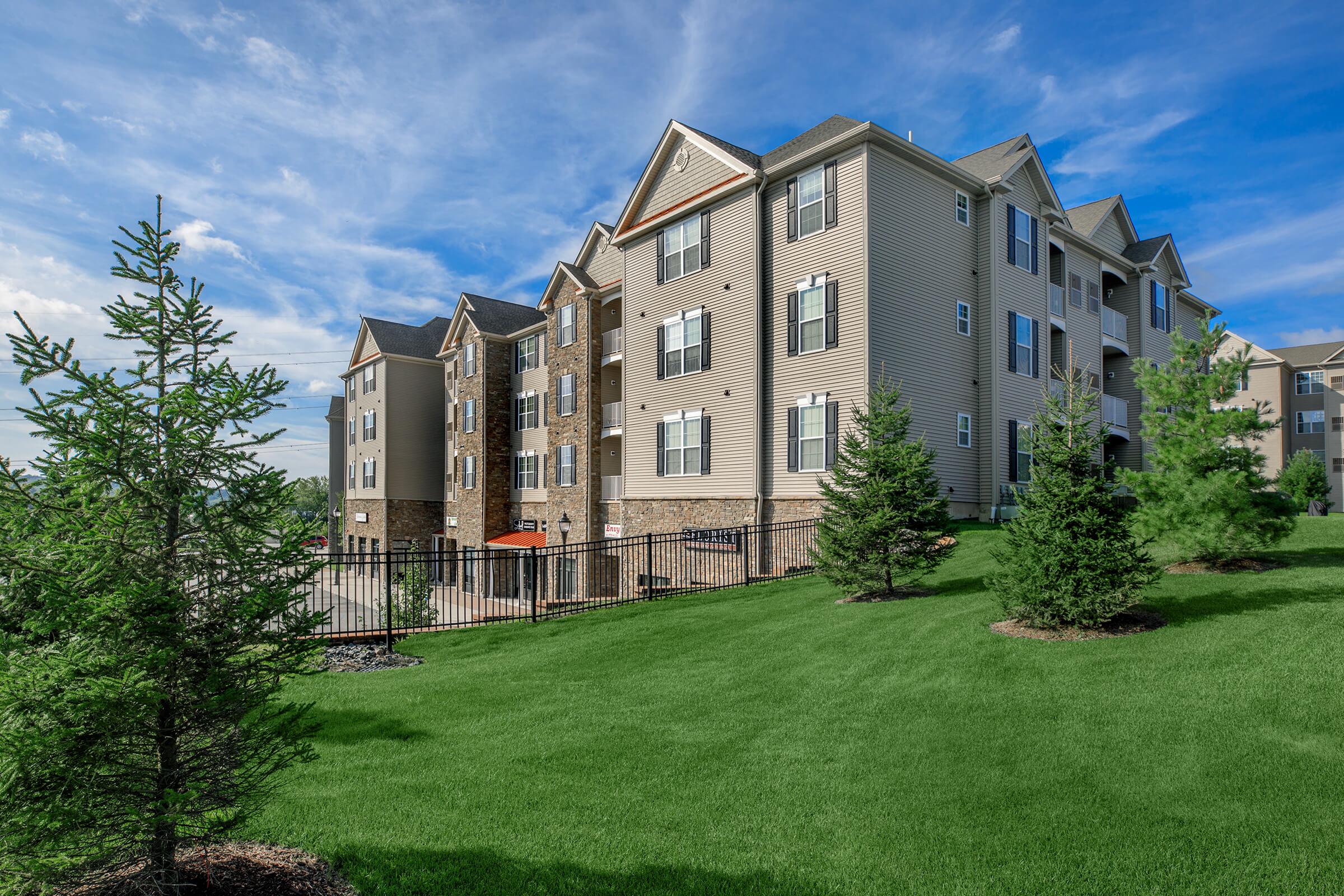A modern apartment building with multiple stories, featuring beige siding and balconies. Lush green grass and small trees frame the property, with a blue sky and scattered clouds in the background, creating a serene and inviting atmosphere.