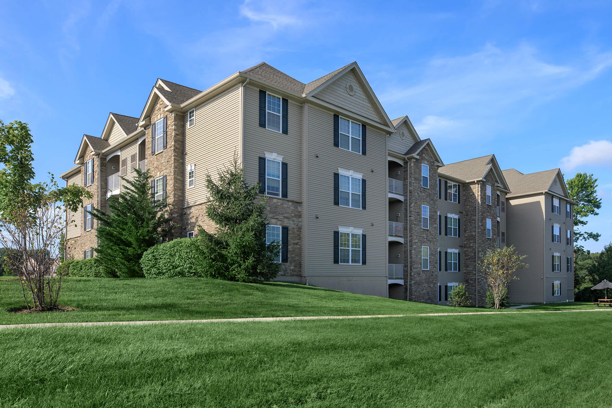 A multi-story residential building with a mix of tan siding and stone accents. The property includes a well-maintained green lawn and shrubs surrounding the building, under a clear blue sky. The architecture features large windows and balconies, creating a welcoming atmosphere.