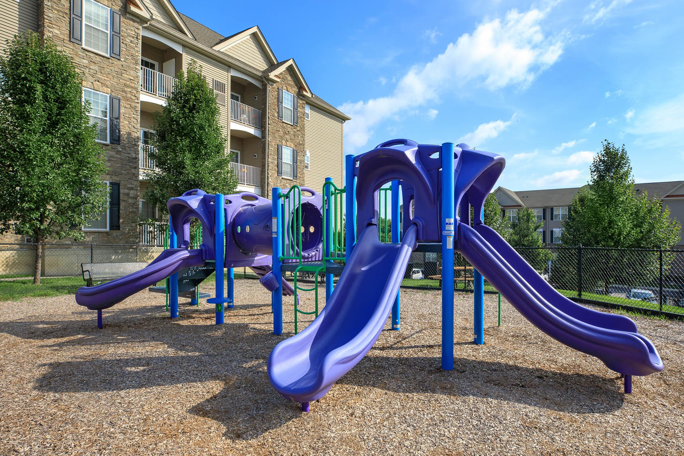 A colorful purple playground structure with slides and climbing features, surrounded by a gravel surface and green trees. In the background, there are multiple apartment buildings under a partly cloudy blue sky.