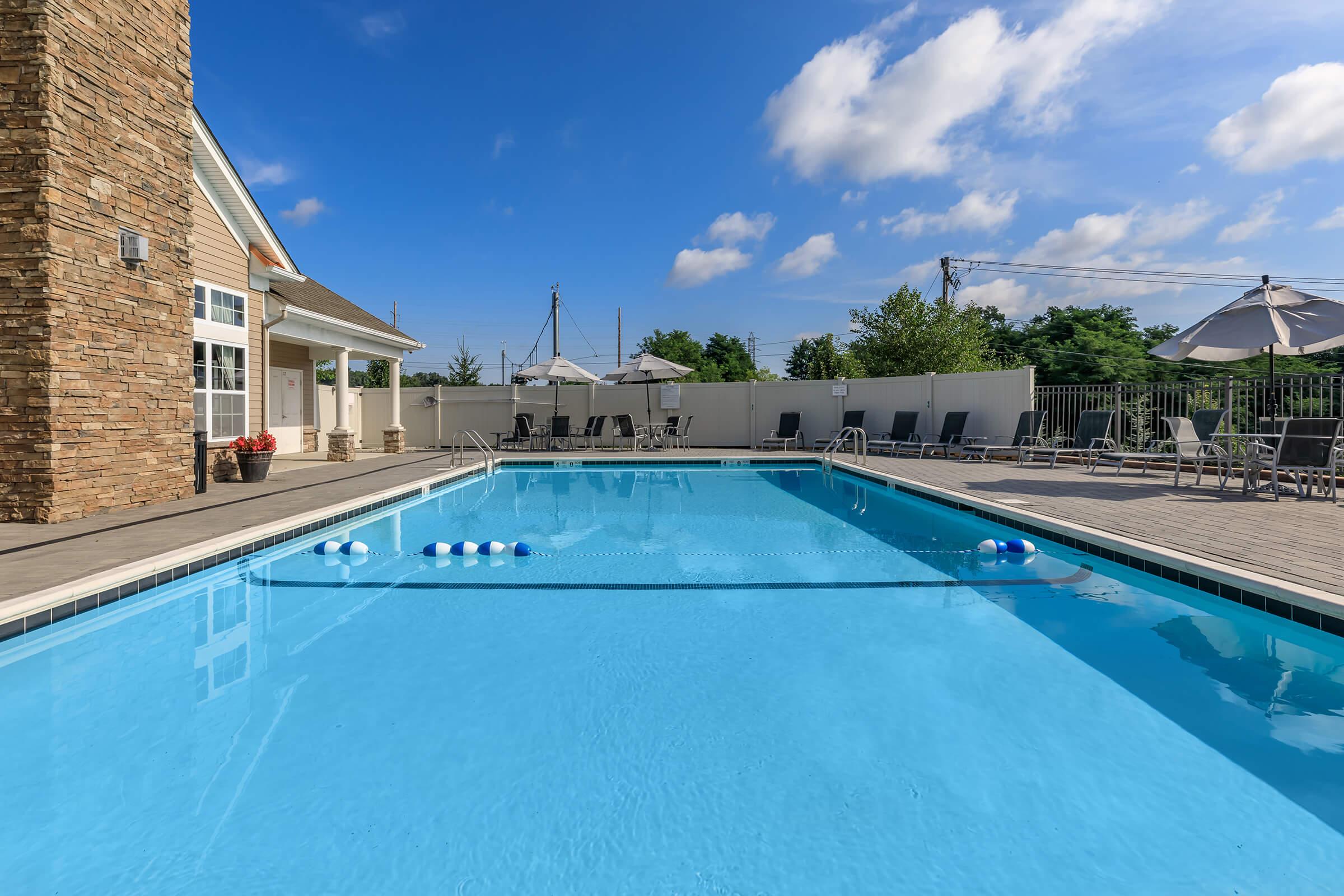 A clear blue swimming pool bordered by a wooden deck, featuring lounge chairs and umbrellas. In the background, a stone-accented building is visible, alongside green trees and a blue sky with scattered clouds. A few floating pool toys rest on the water's surface.