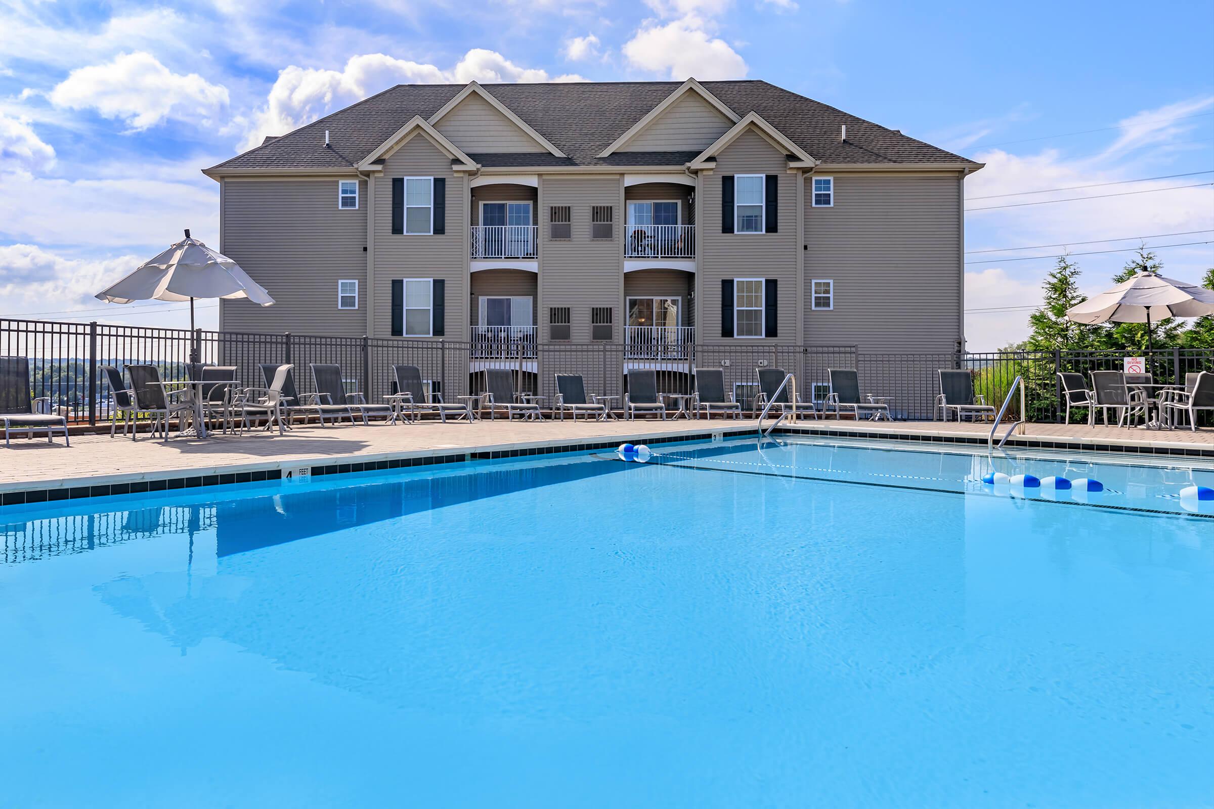 A clear blue swimming pool surrounded by patio furniture and umbrellas, with an apartment building in the background featuring multiple balconies under a bright sky.