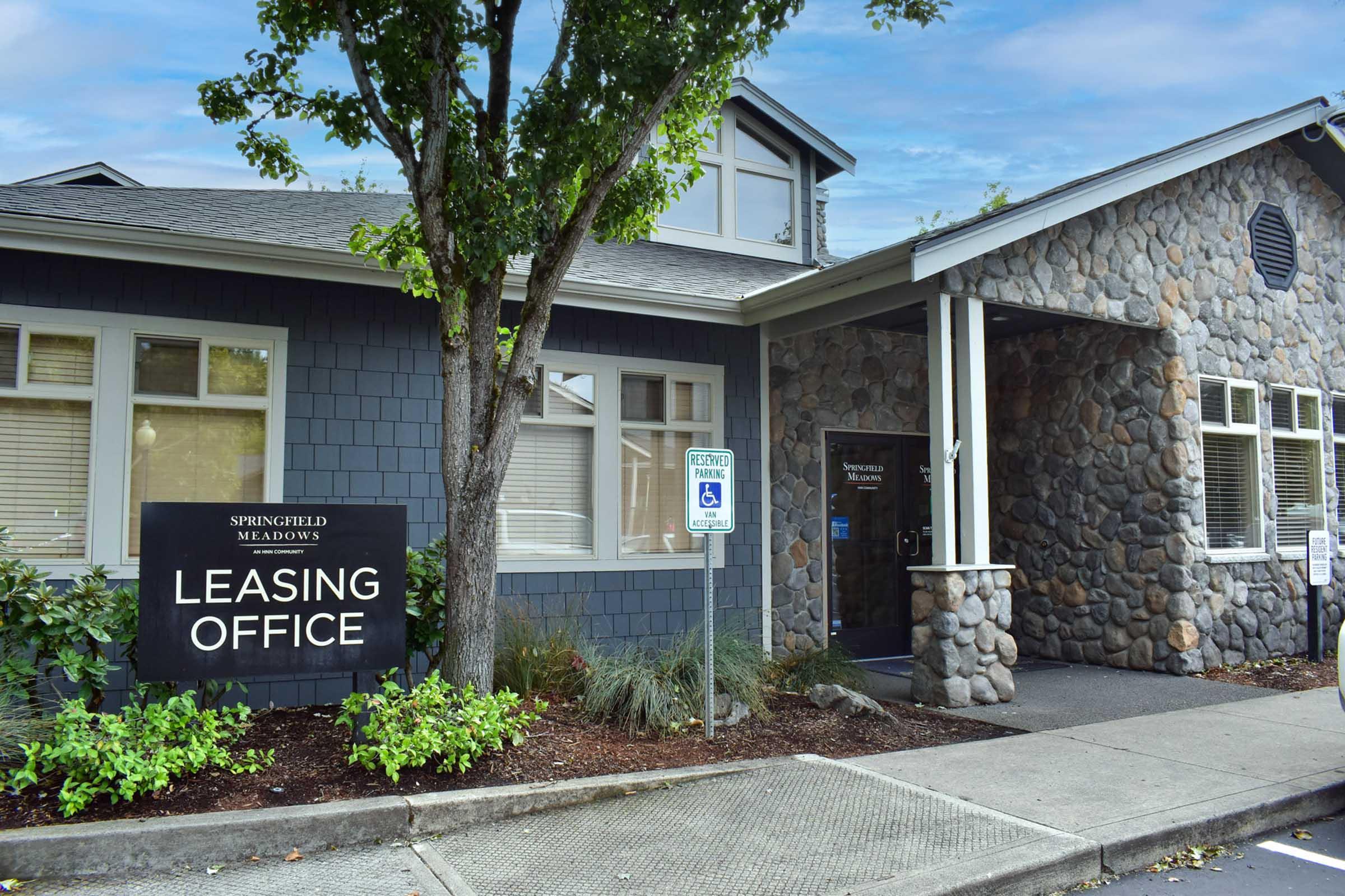 A leasing office building with a stone exterior and a sign that reads "Leasing Office." The structure features large windows, a peaked roof, and is surrounded by greenery. A parking area is visible in front, including a designated handicapped parking space.