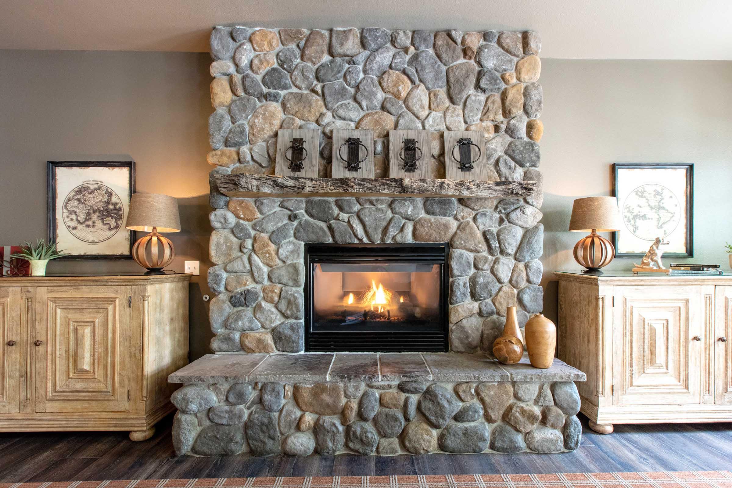 A cozy living room featuring a stone fireplace with a wooden mantel. Above the fireplace, there are four framed artworks. On either side of the fireplace, there are two wooden cabinets with decorative lamps on top. The ambiance is warm and inviting, perfect for relaxation.