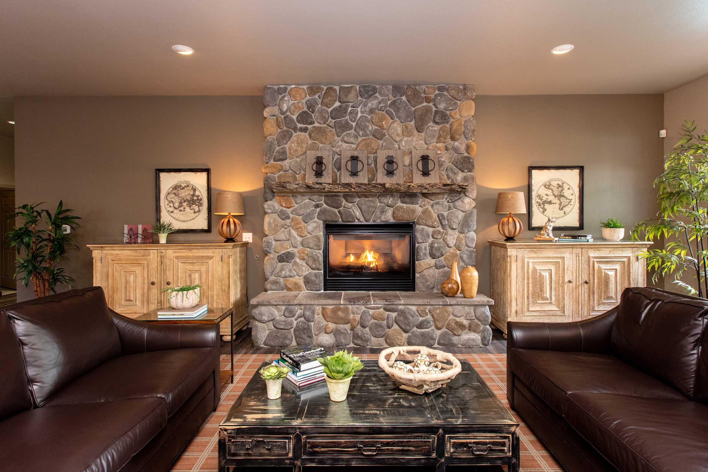 A cozy living room with a stone fireplace at the center. Two dark leather sofas face a wooden coffee table, which features decorative items like plants and a bowl. On either side of the fireplace, there are wooden cabinets with lamps and framed artwork above them, creating a warm and inviting atmosphere.