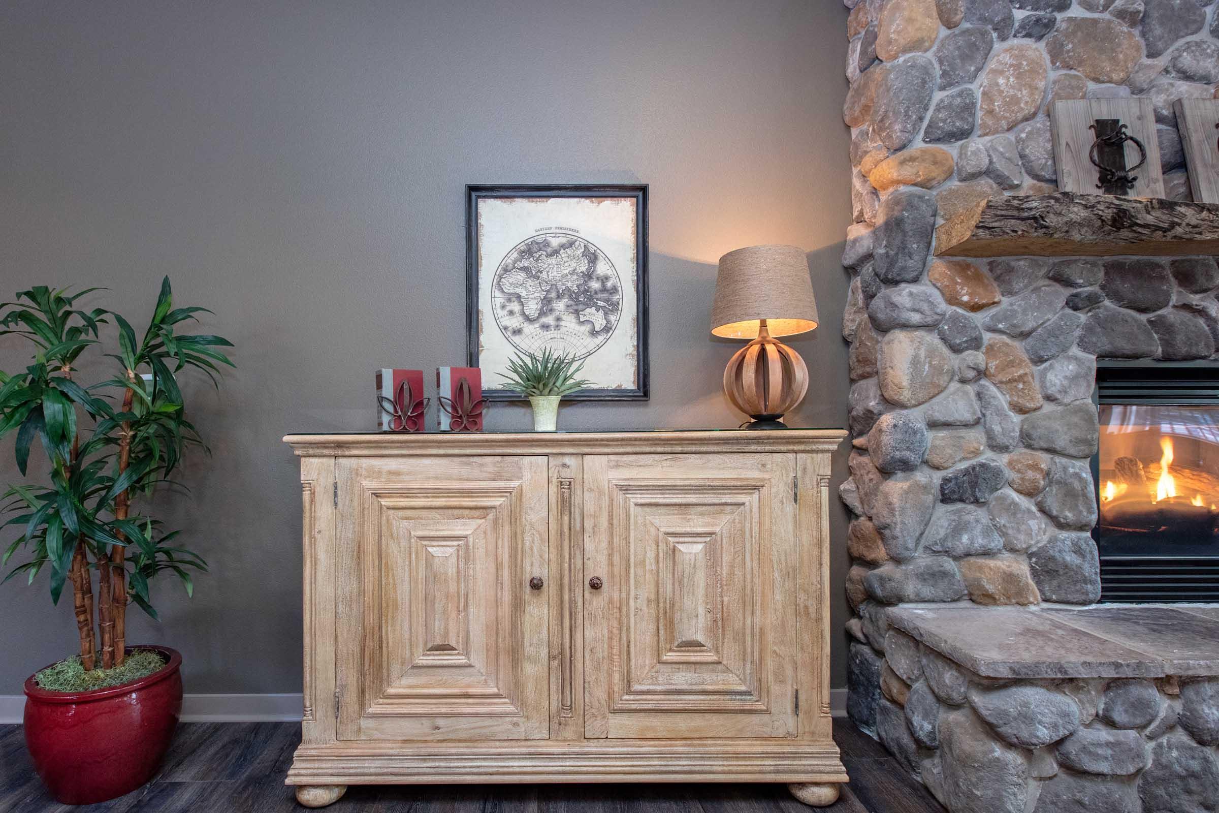 A cozy living space featuring a wooden sideboard with two doors, adorned with decorative items including a potted plant and framed artwork above. A stylish lamp casts warm light next to the sideboard, and a stone fireplace with a visible flame creates a welcoming atmosphere.