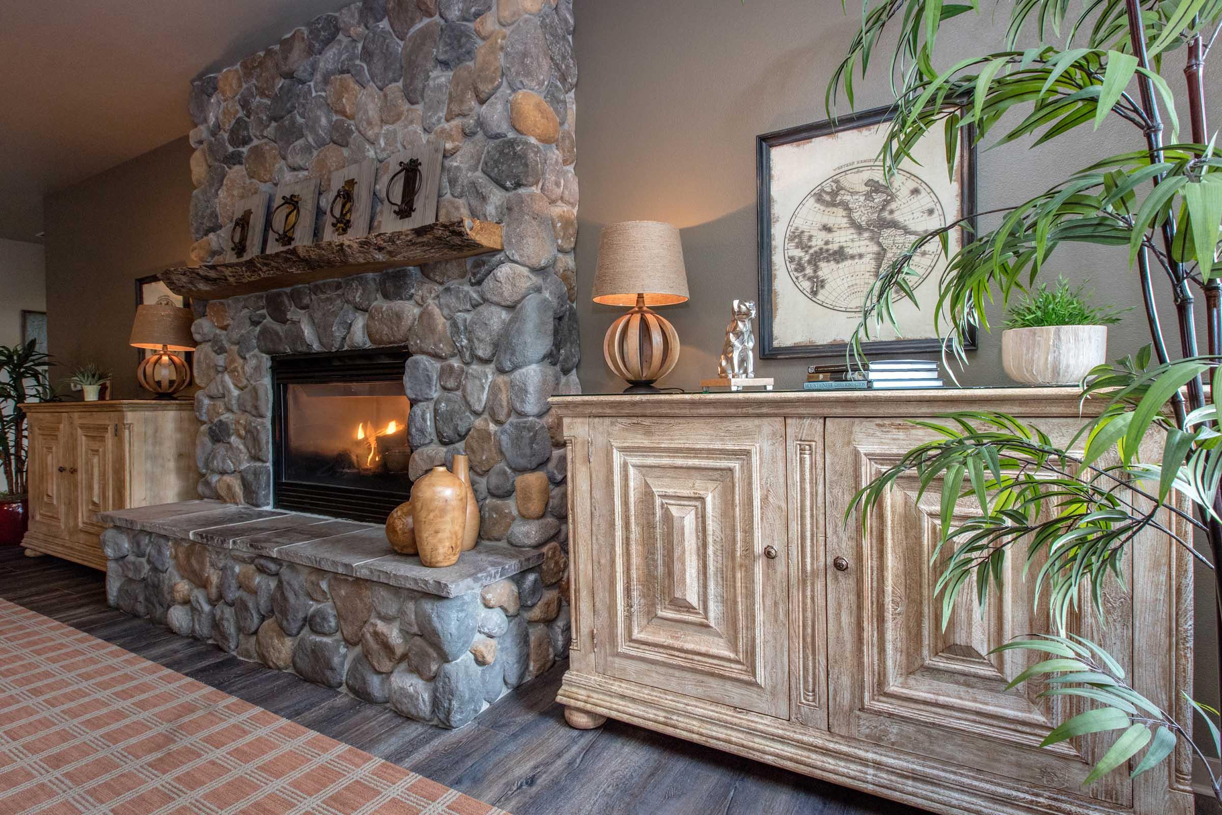 A cozy living room featuring a stone fireplace with a wooden mantel, flanked by two decorative lamps. On either side are rustic cabinets with an earthy color scheme. A potted plant adds a touch of green, while a framed map and books provide homey decor. The floor is covered in neutral-toned area rugs.