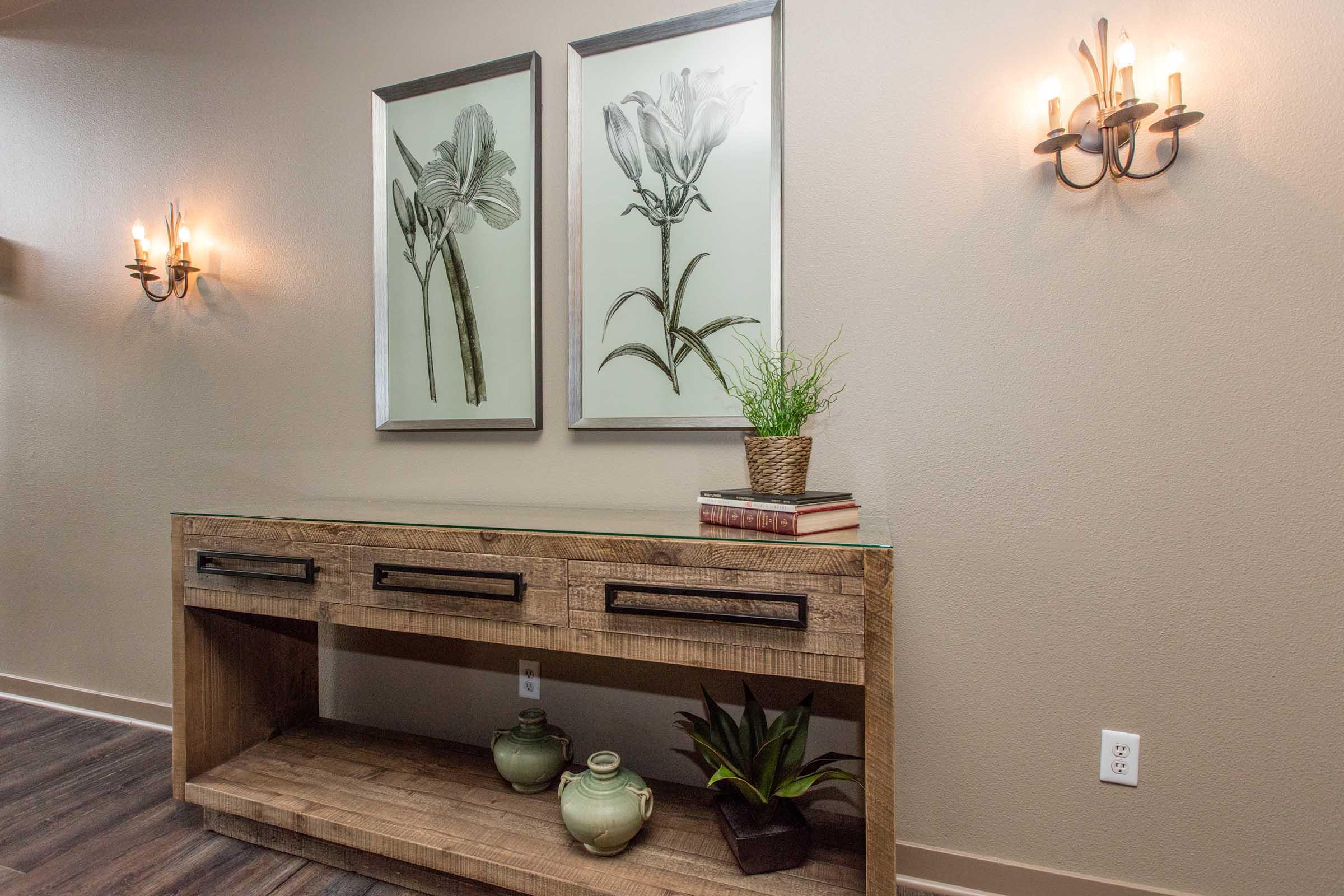 A wooden console table with three drawers, positioned against a wall. Above the table, there are two framed botanical prints of flowers. To the left, there are two wall sconces providing warm lighting. On the table, a decorative green plant and two light green vases add to the decor.