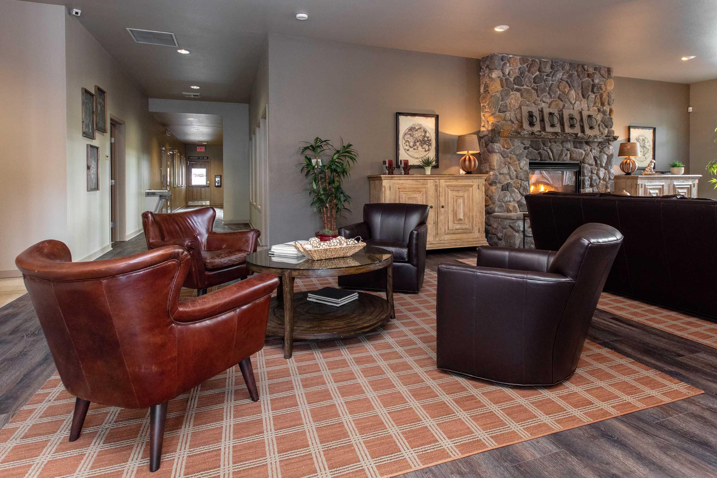 A cozy lounge area featuring two brown leather armchairs and a round table with decorative items. The space has a stone fireplace in the background, surrounded by wooden furniture and plants, with a large rug on the floor, creating a warm and inviting atmosphere.