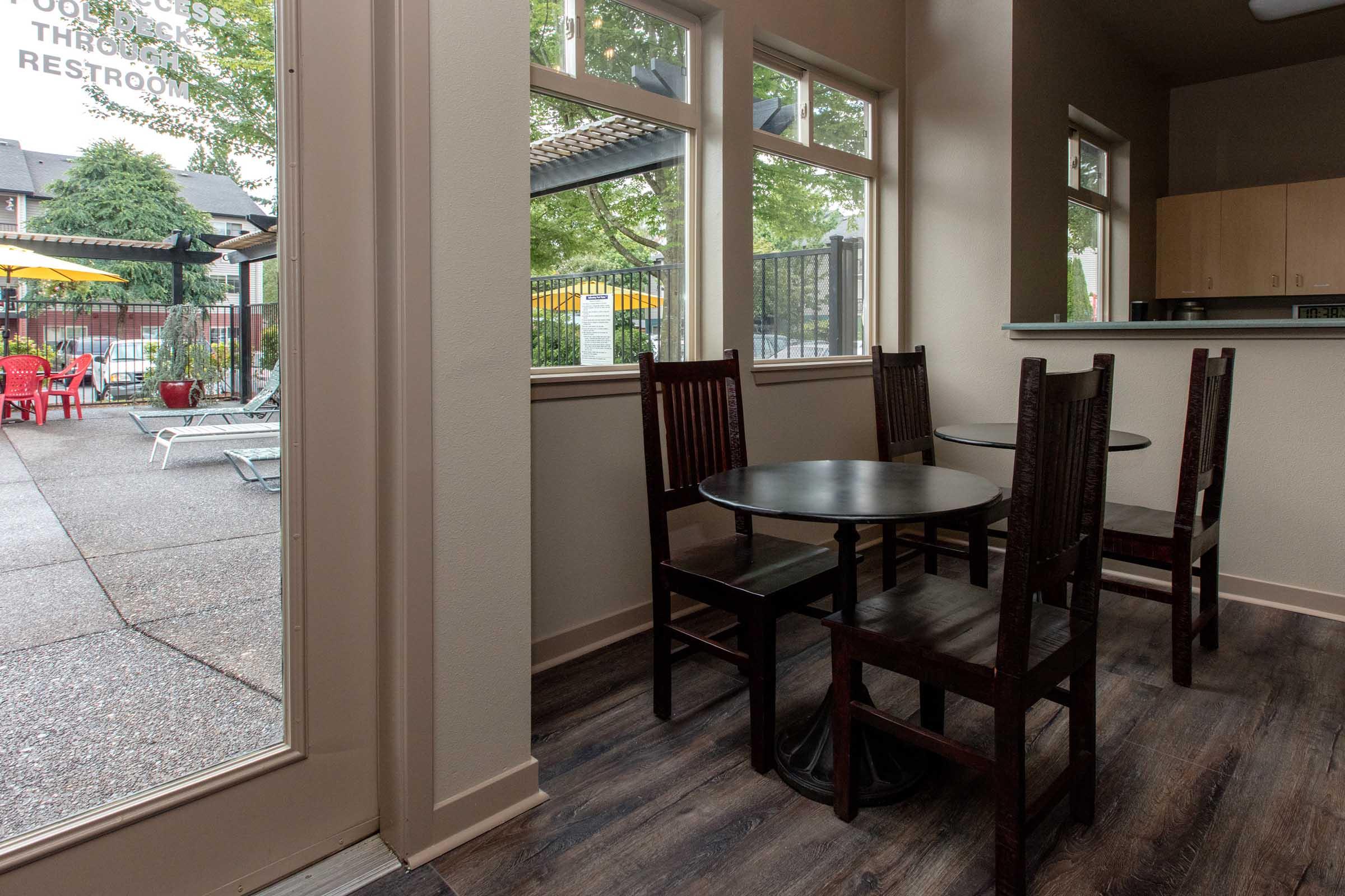 A small indoor seating area with a round table and four wooden chairs, illuminated by natural light from large windows. Outside, a view of a pool area with lounge chairs and colorful umbrellas can be seen, creating a relaxed and inviting atmosphere.
