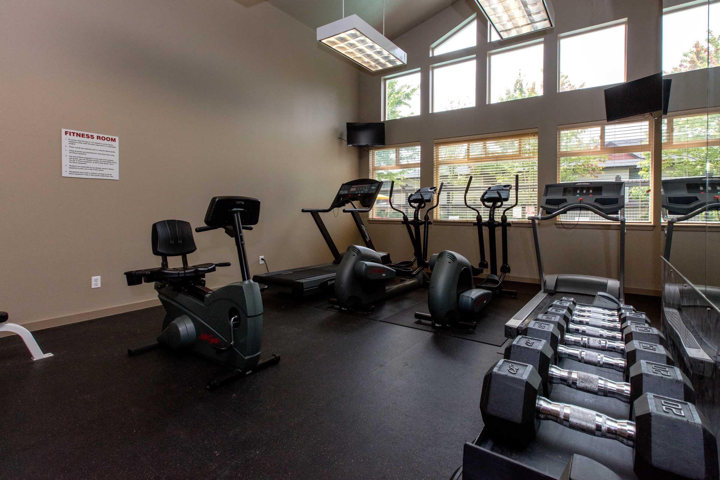 A fitness room featuring exercise equipment including stationary bikes, treadmills, and dumbbells, with large windows allowing natural light. The space is designed for workout activities, with wall mirrors and a fitness information sign displayed.