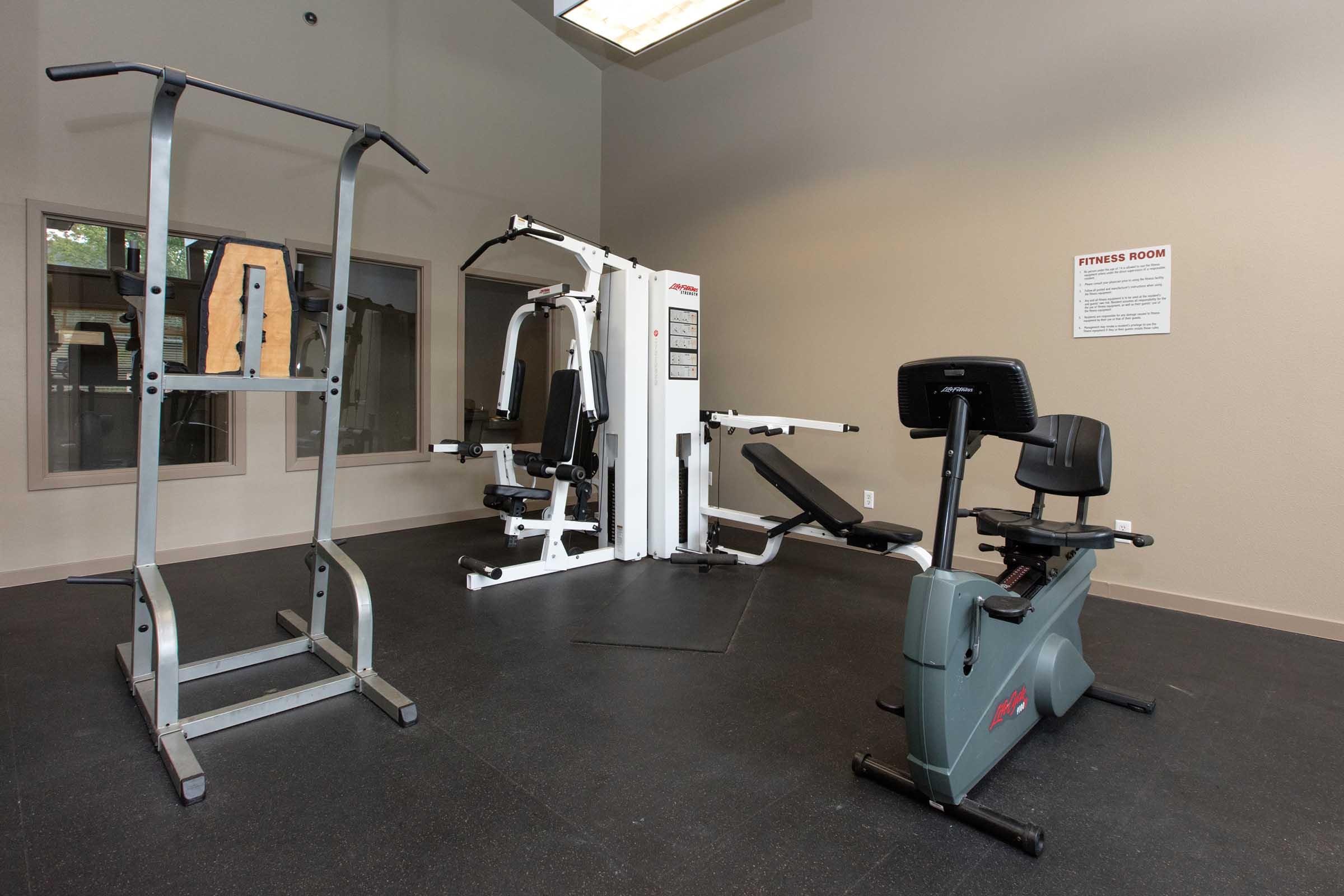 A fitness room featuring a rowing machine, multi-exercise equipment, and a weight lifting station. The space is well-lit with windows and includes informational signage about the fitness area. The flooring is made of black rubber material for safety and comfort during workouts.