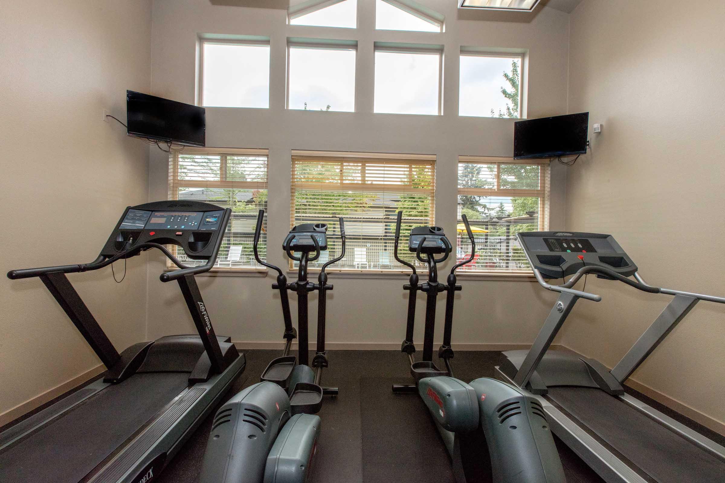 A modern fitness room featuring two treadmills, two elliptical machines, and large windows allowing natural light. The space is equipped with two wall-mounted TVs and has a clean, well-lit environment designed for exercise.