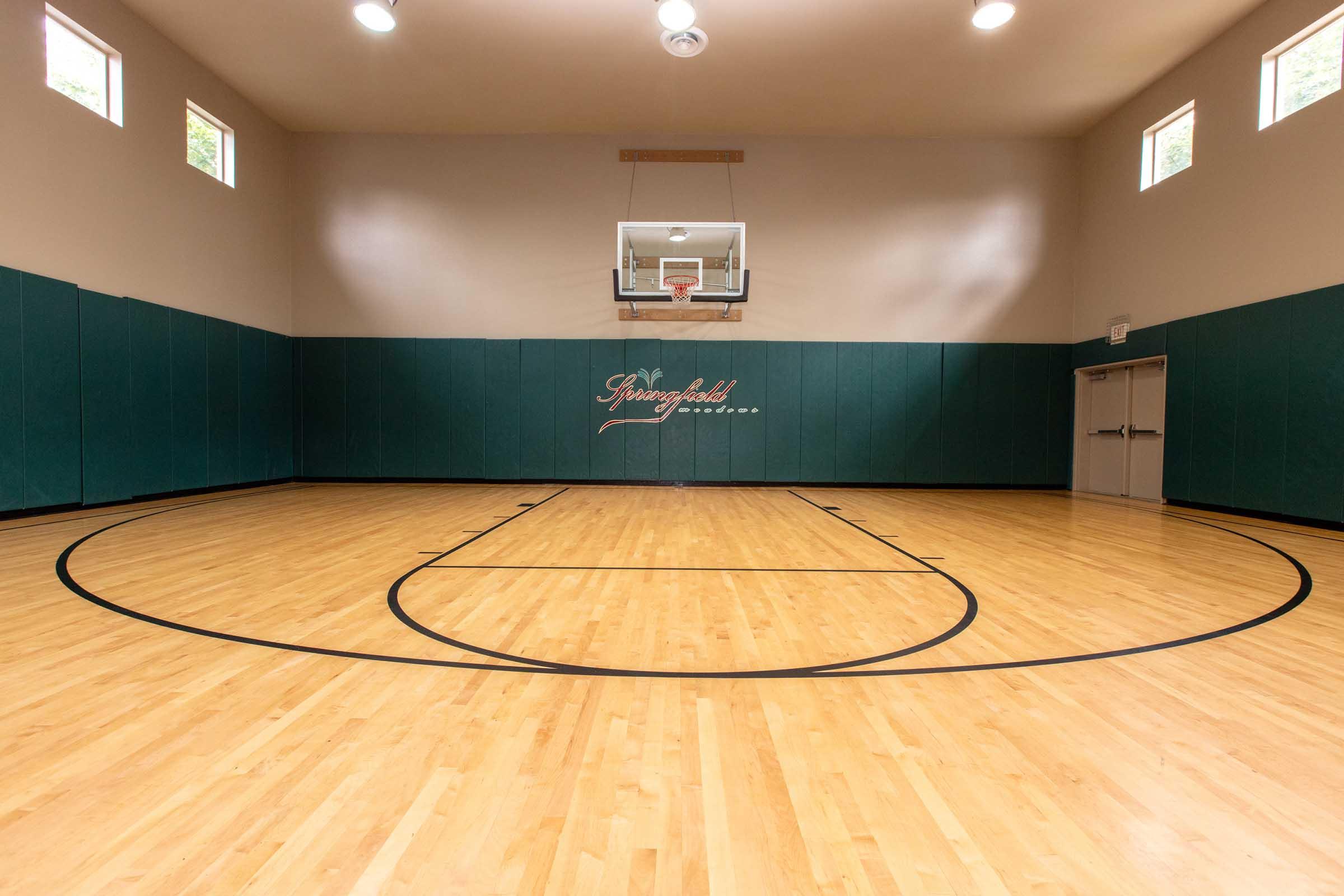 An indoor basketball court with polished wooden flooring, black basketball court lines, and a hoop mounted on the wall. The walls are painted green, with small windows near the ceiling allowing natural light. The court is empty, creating a spacious and inviting atmosphere for play.