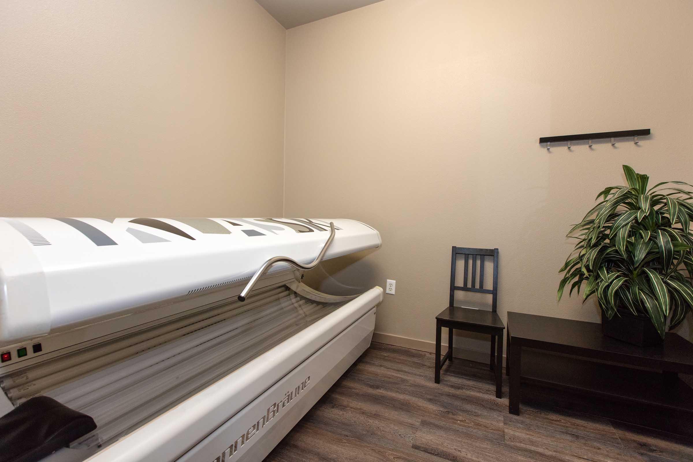 A small, well-lit room featuring a futuristic tanning bed with a sleek design. A single chair is positioned nearby, along with a small table. There's a green potted plant in the corner, adding a touch of decor to the space. The walls are painted a neutral color, giving the room a clean and modern look.