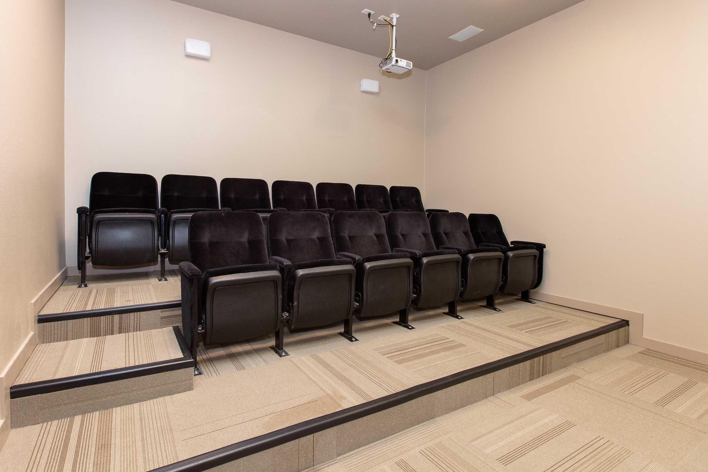 A small, modern theater room featuring several black upholstered chairs arranged in a tiered formation. There is a projector mounted on the ceiling, and the floor is covered with light beige carpeting. The room has neutral-colored walls and a clean, minimalistic design, creating a private viewing space.