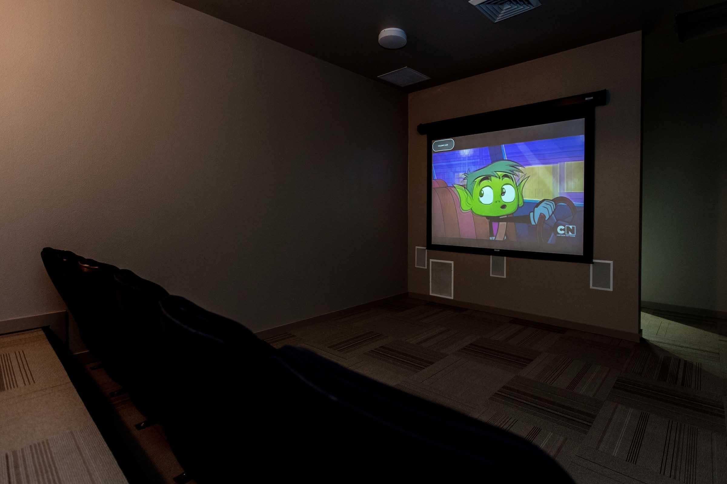 A cozy home theater setting with several empty seats facing a wall-mounted projector screen. The screen displays an animated character with green skin and big eyes, looking surprised or excited. The room has muted lighting and neutral-colored walls, creating an intimate viewing atmosphere.