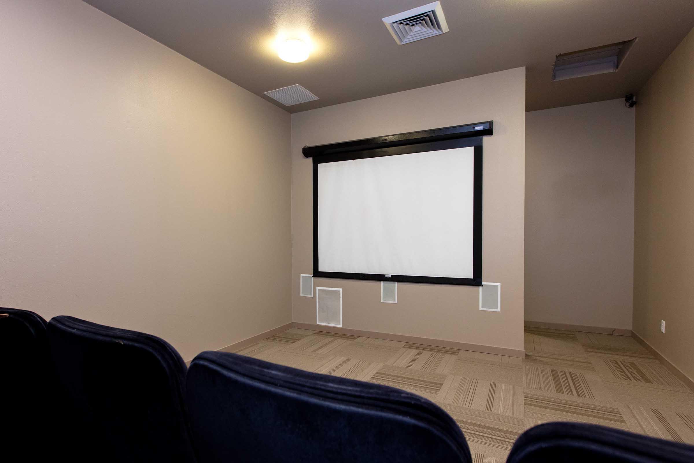 A small, dimly lit room designed for presentations or screenings, featuring a wall-mounted projection screen at the front. There are several empty black chairs arranged in rows facing the screen, with beige walls and a carpeted floor. The space appears modern and minimalistic.