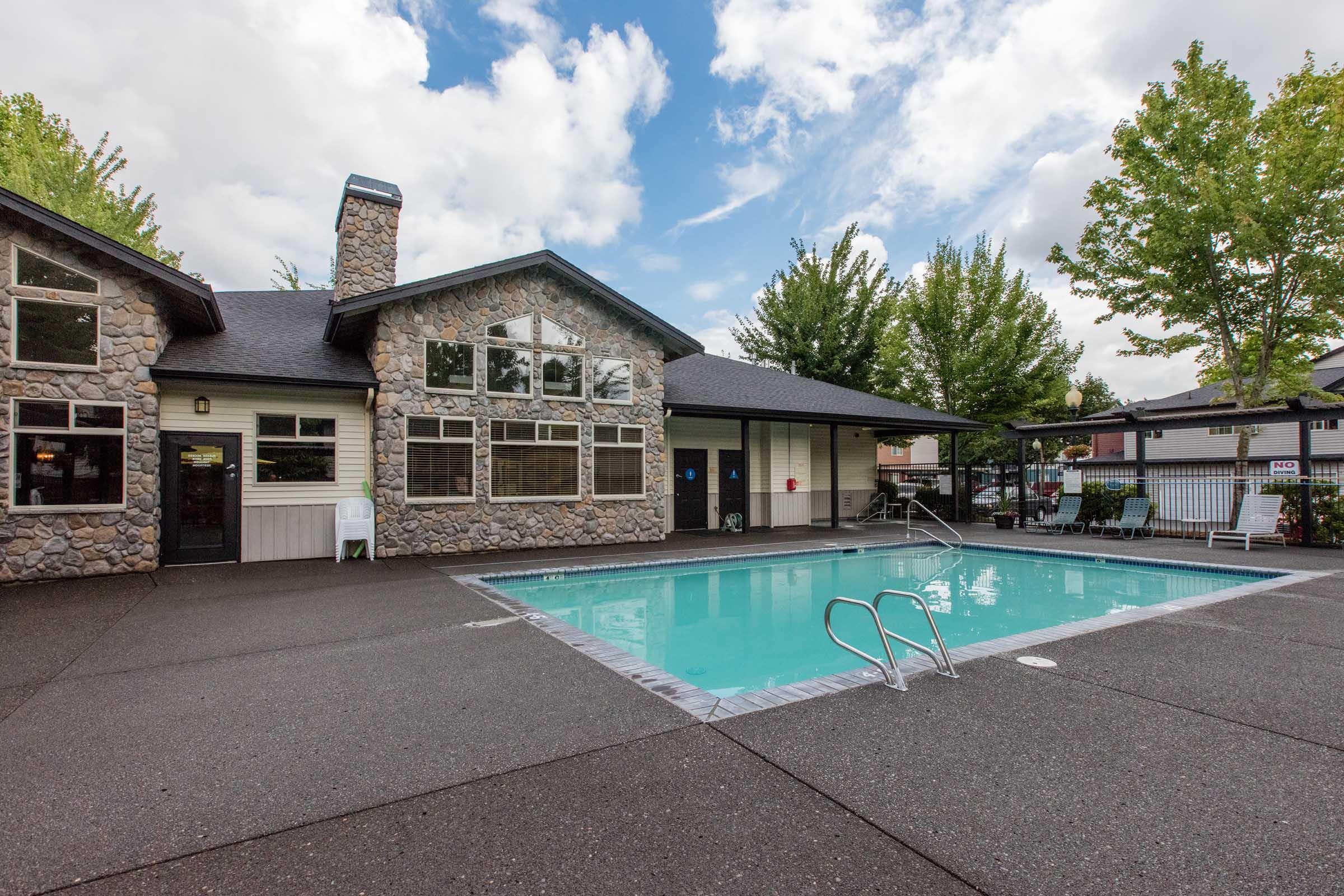 A swimming pool surrounded by a stone building with large windows, set in a landscaped area with trees. The clear blue water reflects the cloudy sky, and there are poolside chairs and a diving board. The scene conveys a peaceful, recreational atmosphere.