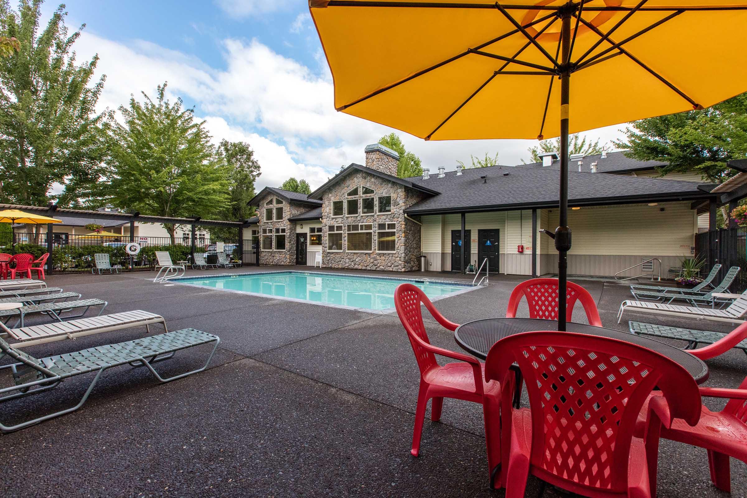 A bright outdoor pool area featuring a clear blue pool surrounded by lounge chairs and tables. Yellow umbrellas provide shade, and the backdrop includes a modern stone building with large windows and trees. The atmosphere is inviting and relaxed, ideal for leisure activities.