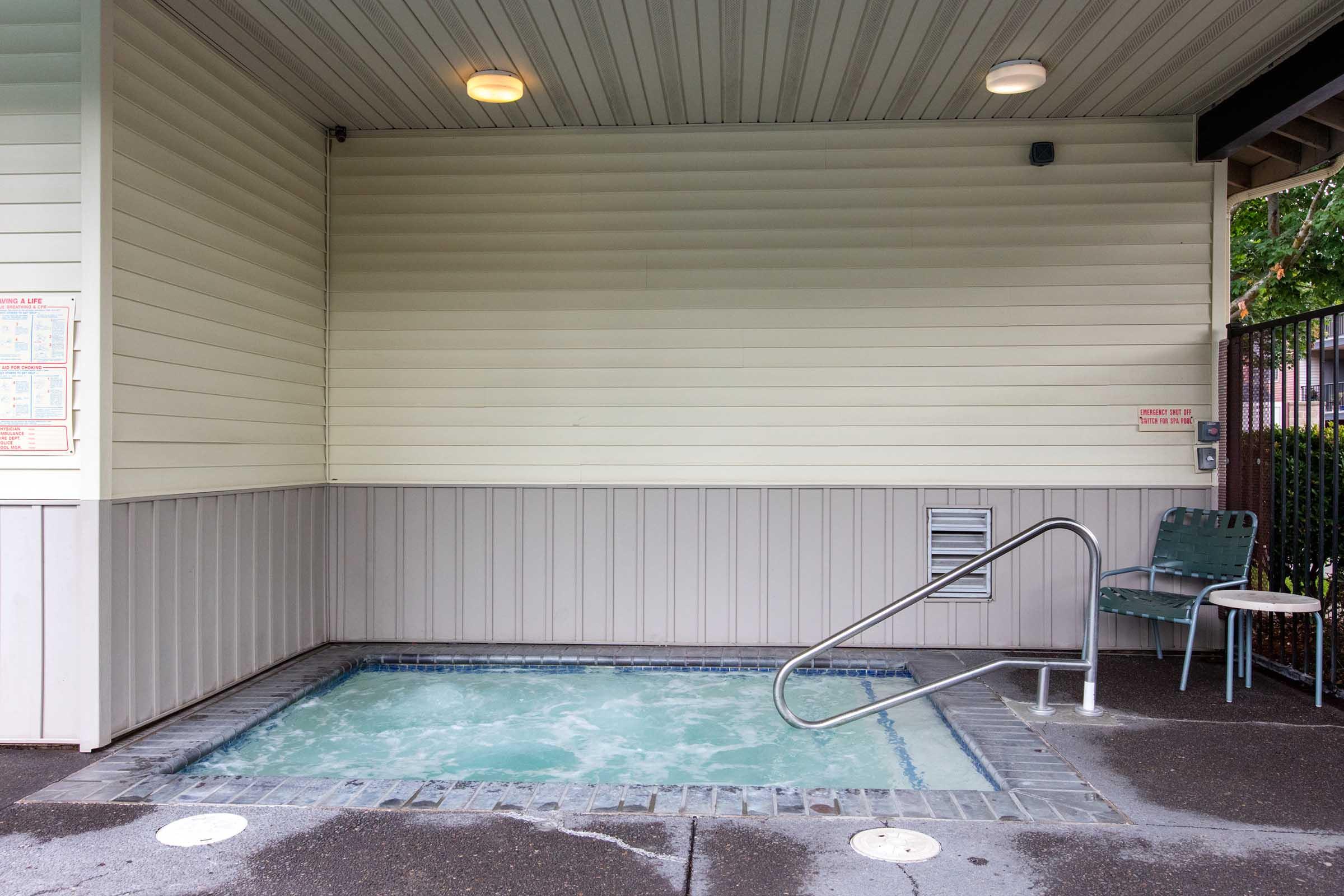A small, outdoor hot tub with bubbling water is situated against a light-colored wall. A metal handrail leads into the tub, and there is a green chair nearby. The area is clean and well-maintained, with overhead lighting and a gated entrance visible in the background.